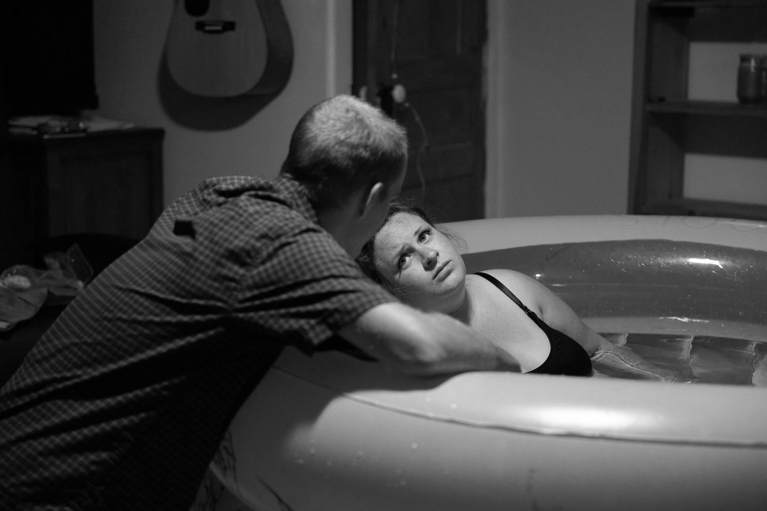 Woman laying in a birthing pool gets support during water birth in black and white photograph.