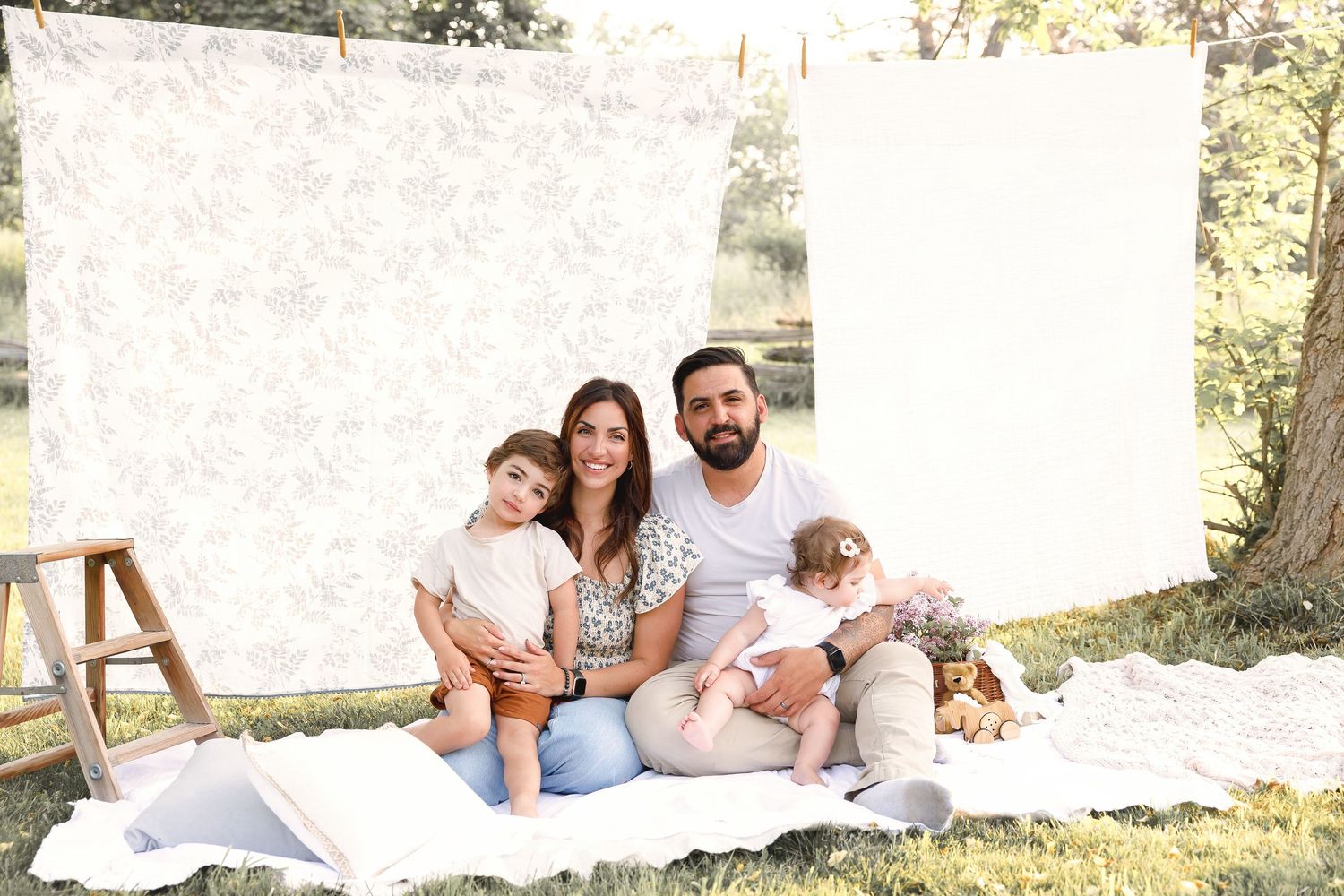 A family sits together on a white blanket outdoors against a light backdrop on a sunny day with natural surroundings.