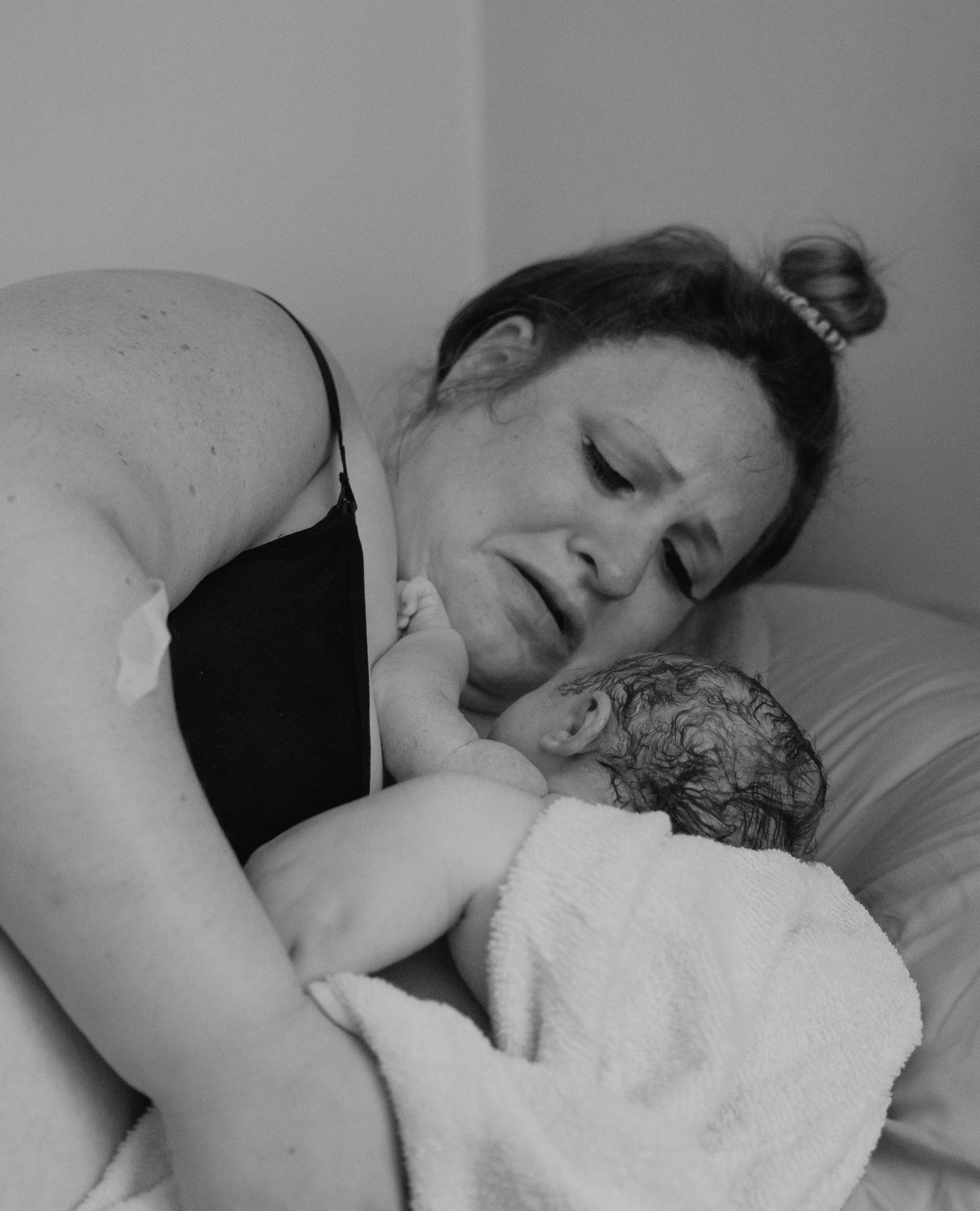 A tender black and white photograph captures an intimate postpartum moment between parent and newborn in bed.