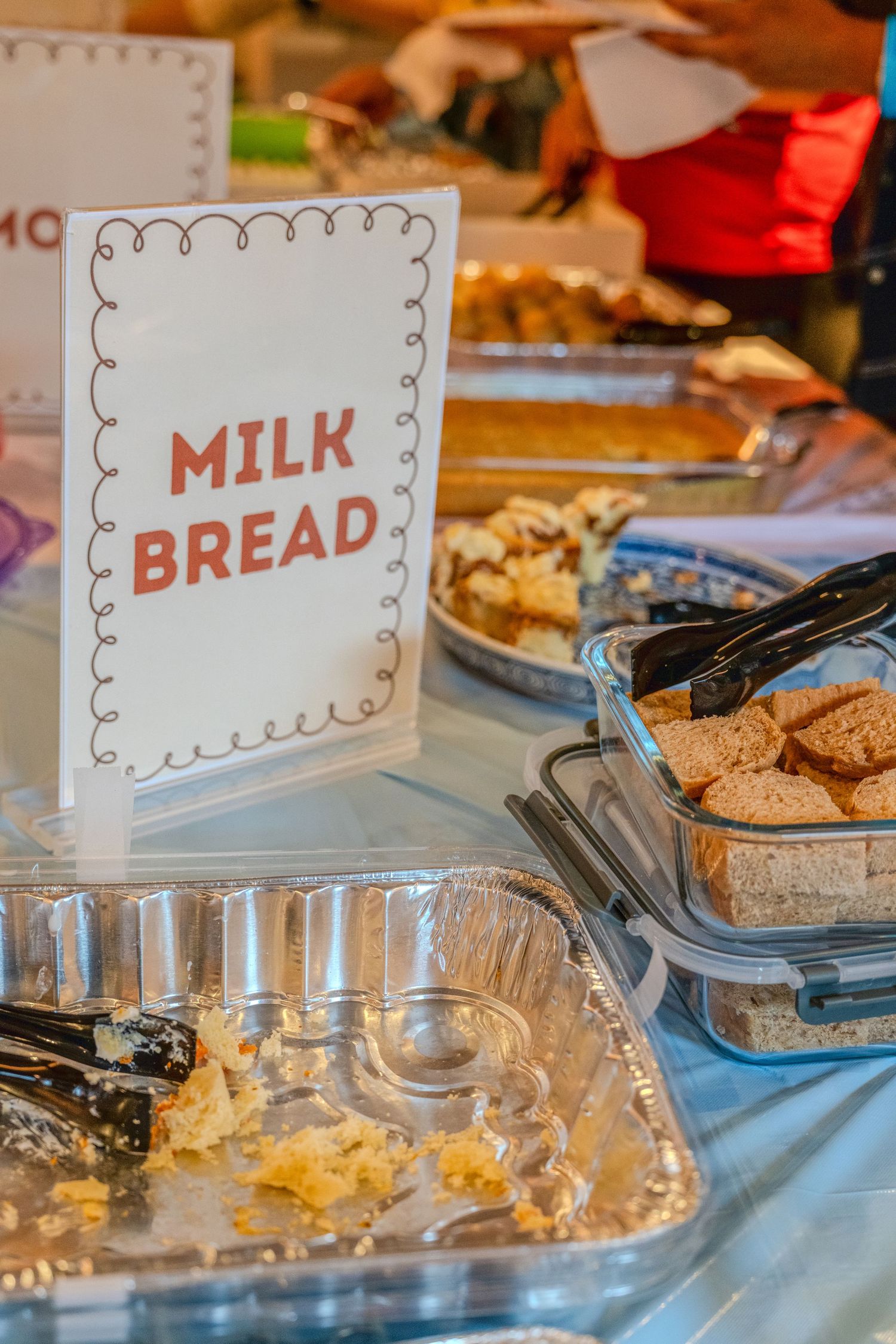 A serving table displays a 'Milk Bread' sign with aluminum trays of food and cookies at what appears to be a buffet setup.
