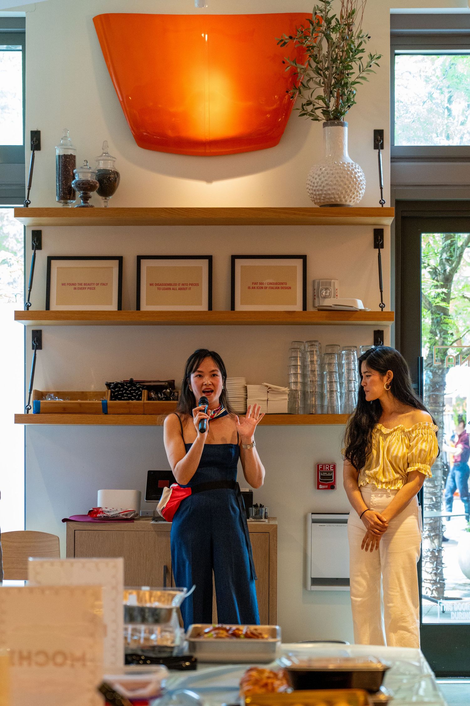 Two people stand in front of wooden shelves with a large orange wall light fixture in a retail boutique interior.