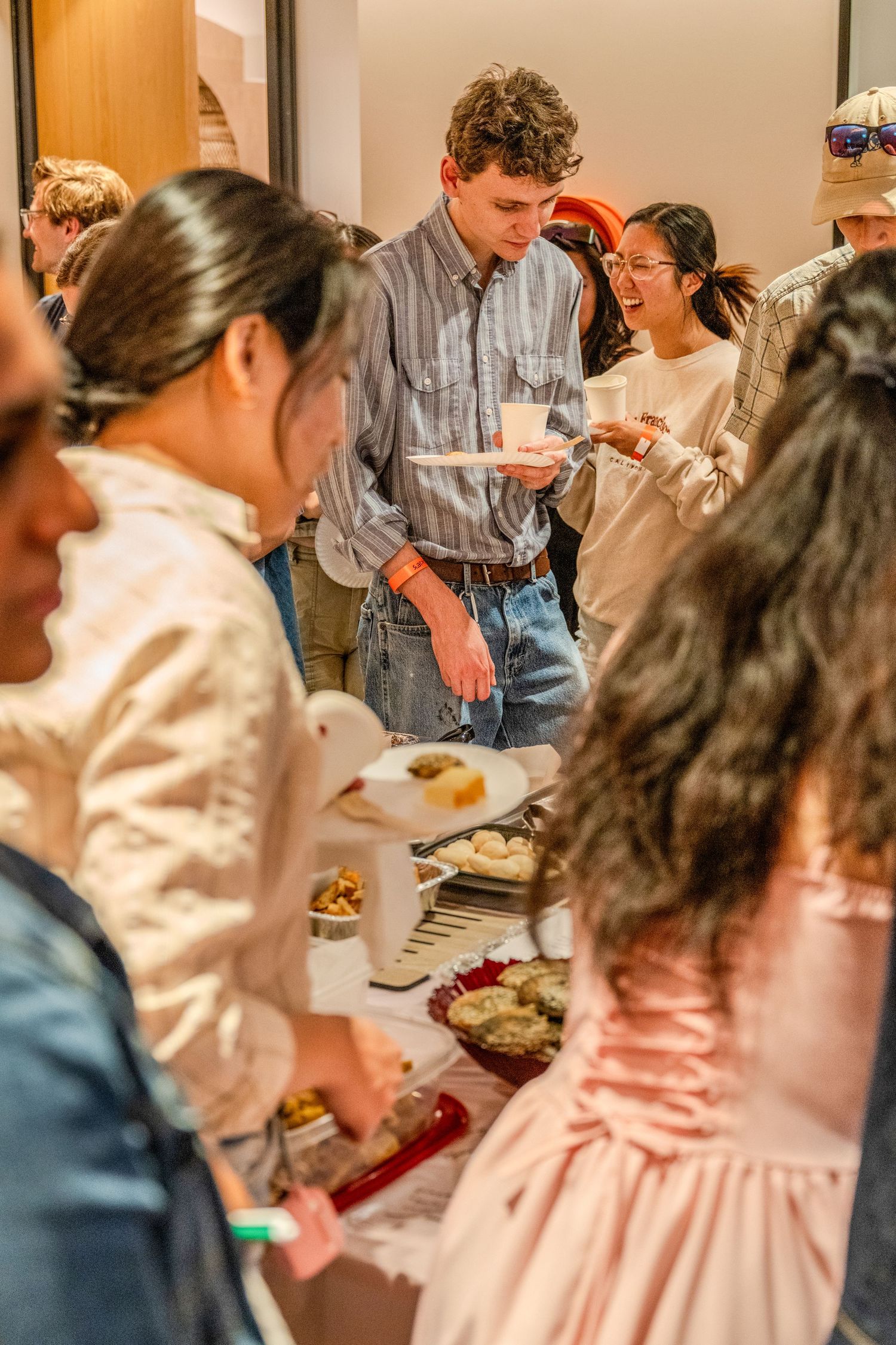 People gathered around a buffet table at a social event, serving themselves food and drinks in a warm-lit indoor setting.