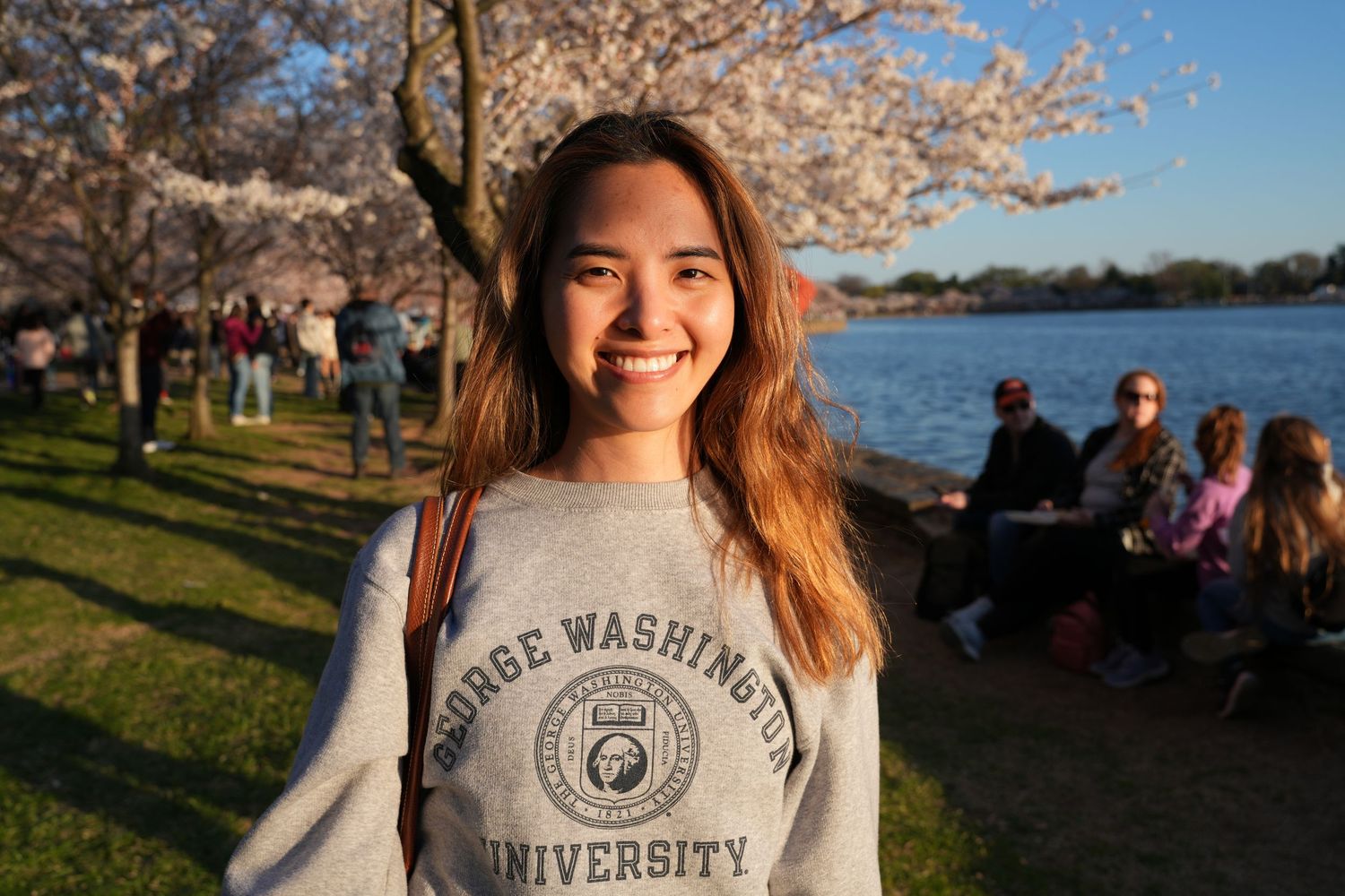 A student wearing a George Washington University sweatshirt smiles near cherry blossoms at the Tidal Basin.