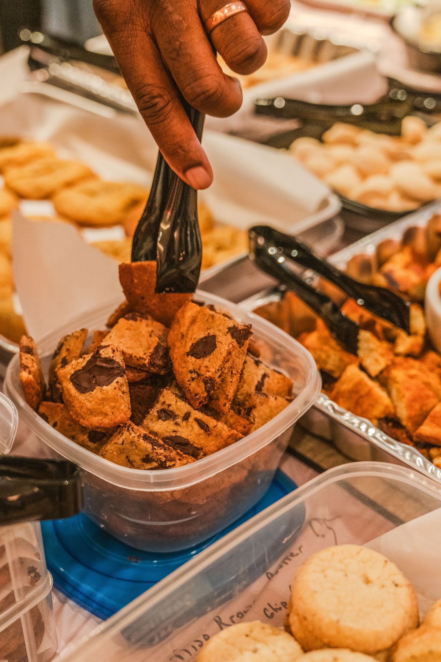 Hand using tongs to serve freshly baked chocolate chip cookies from a plastic container at a dessert buffet display.