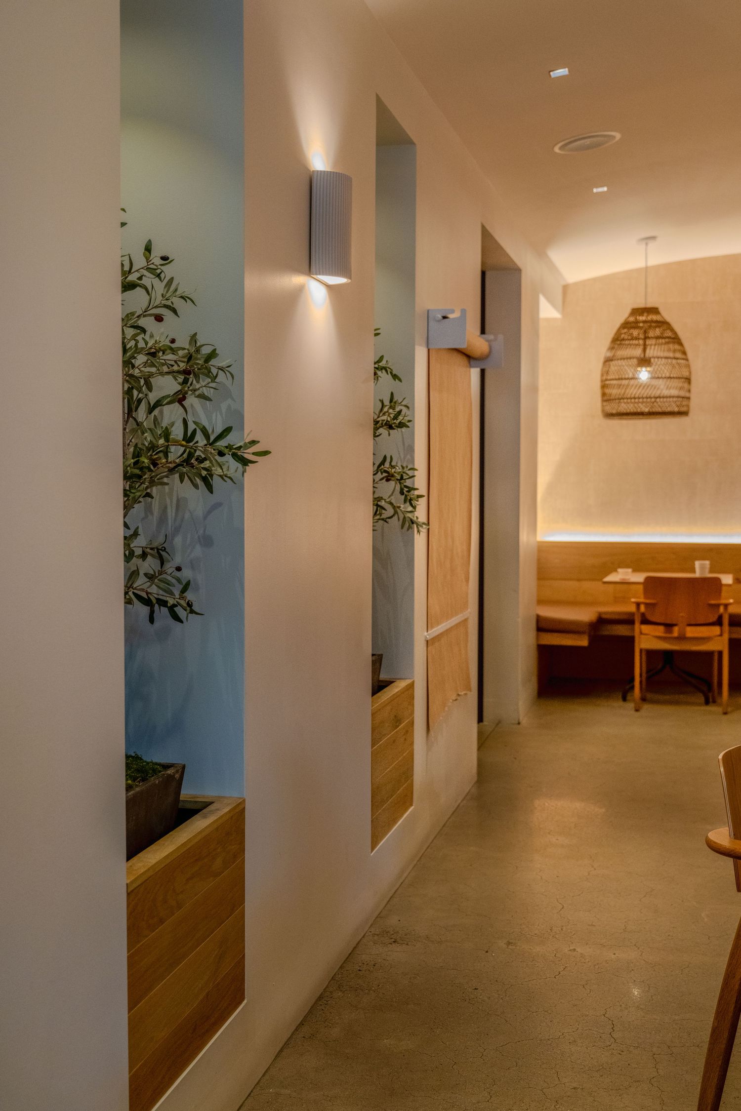 Modern restaurant interior with warm lighting, wooden planters, and a woven pendant lamp along a minimalist hallway.
