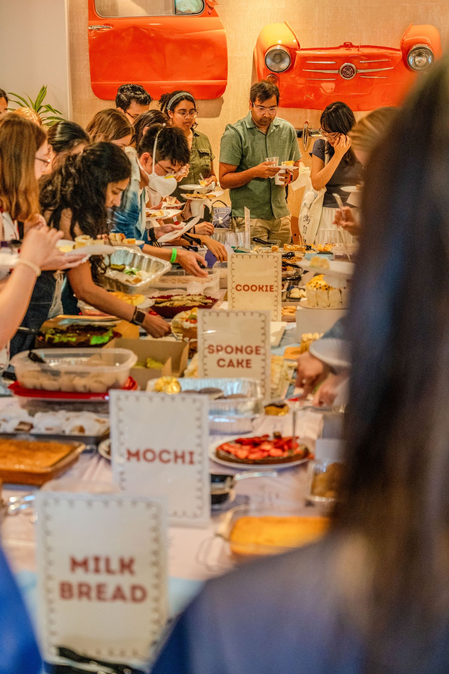 A buffet table with labeled desserts including milk bread, mochi, and sponge cake while people gather to serve themselves.