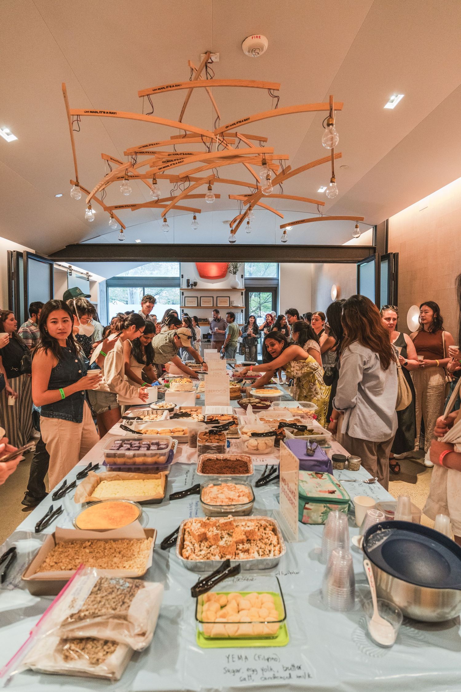 A crowded buffet table with various dishes and desserts at a social gathering under an orange geometric chandelier.