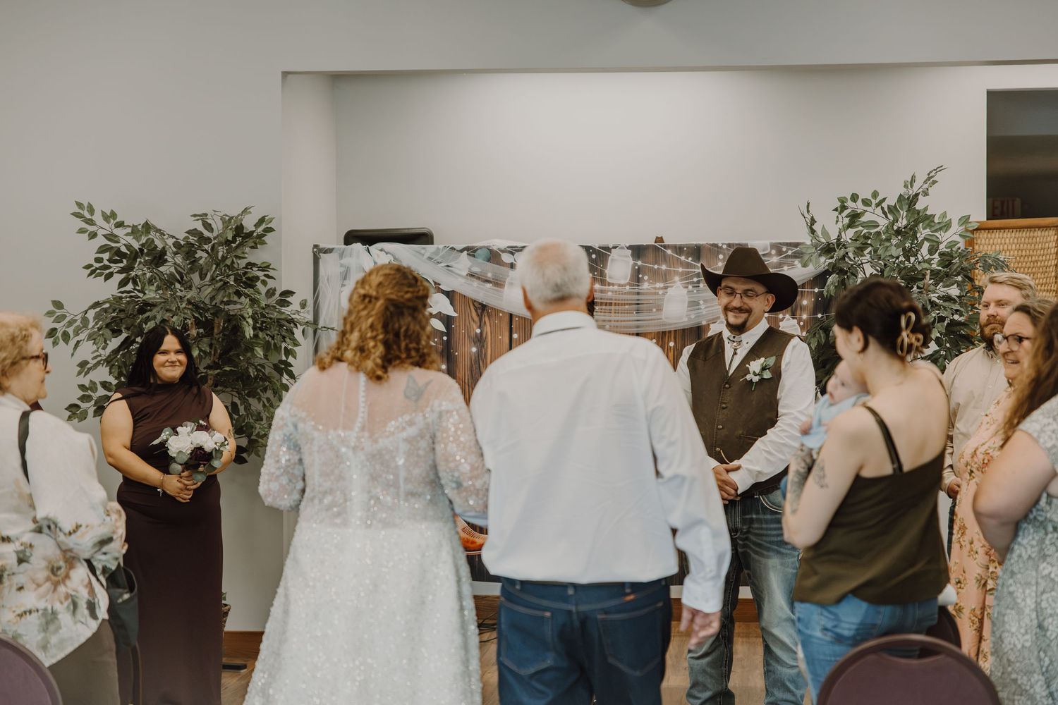 A bride and groom exchange vows at an indoor wedding ceremony surrounded by guests and greenery decorations.