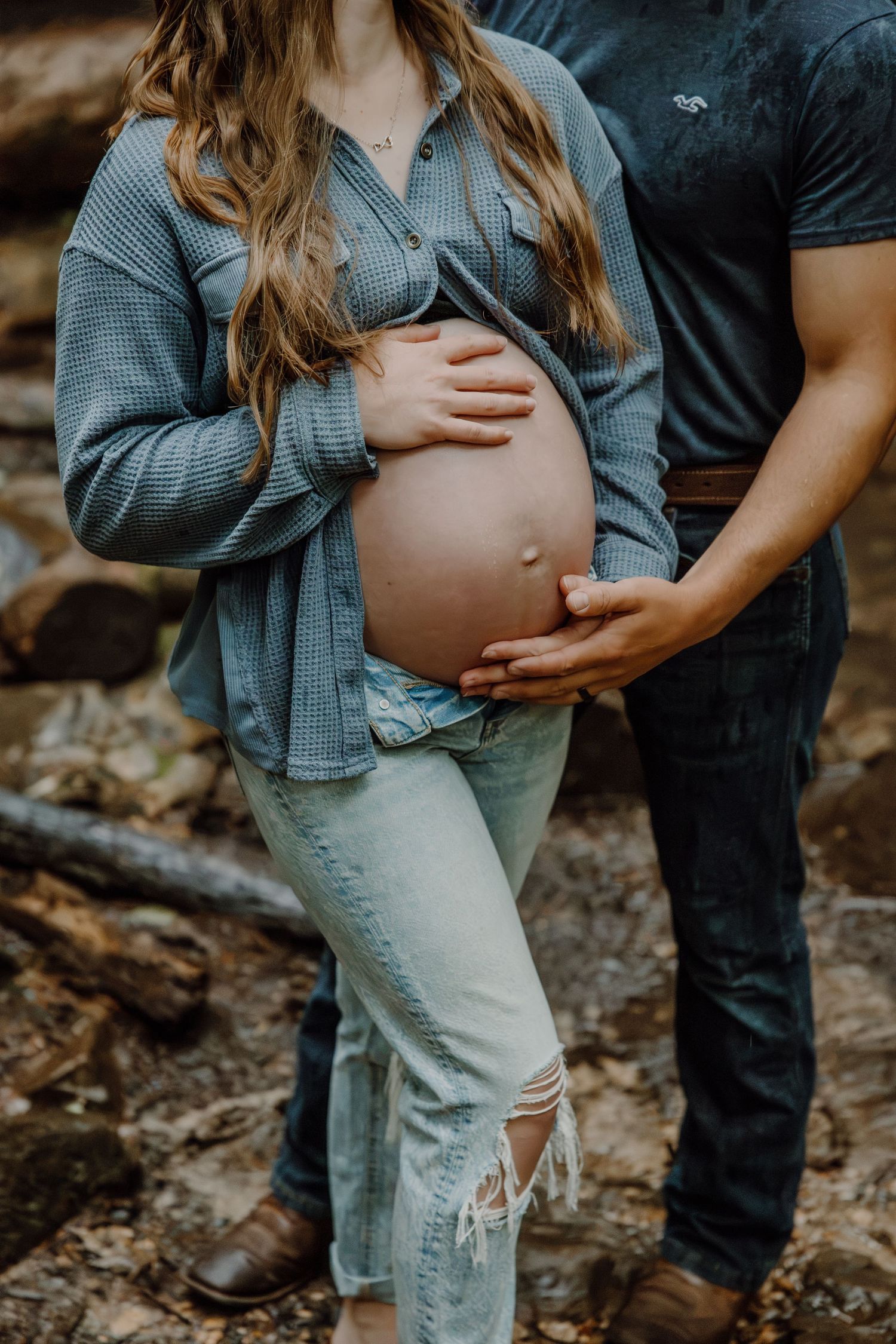 Couple posing for outdoor maternity photo shoot, showing pregnant belly with hands cradling bump.