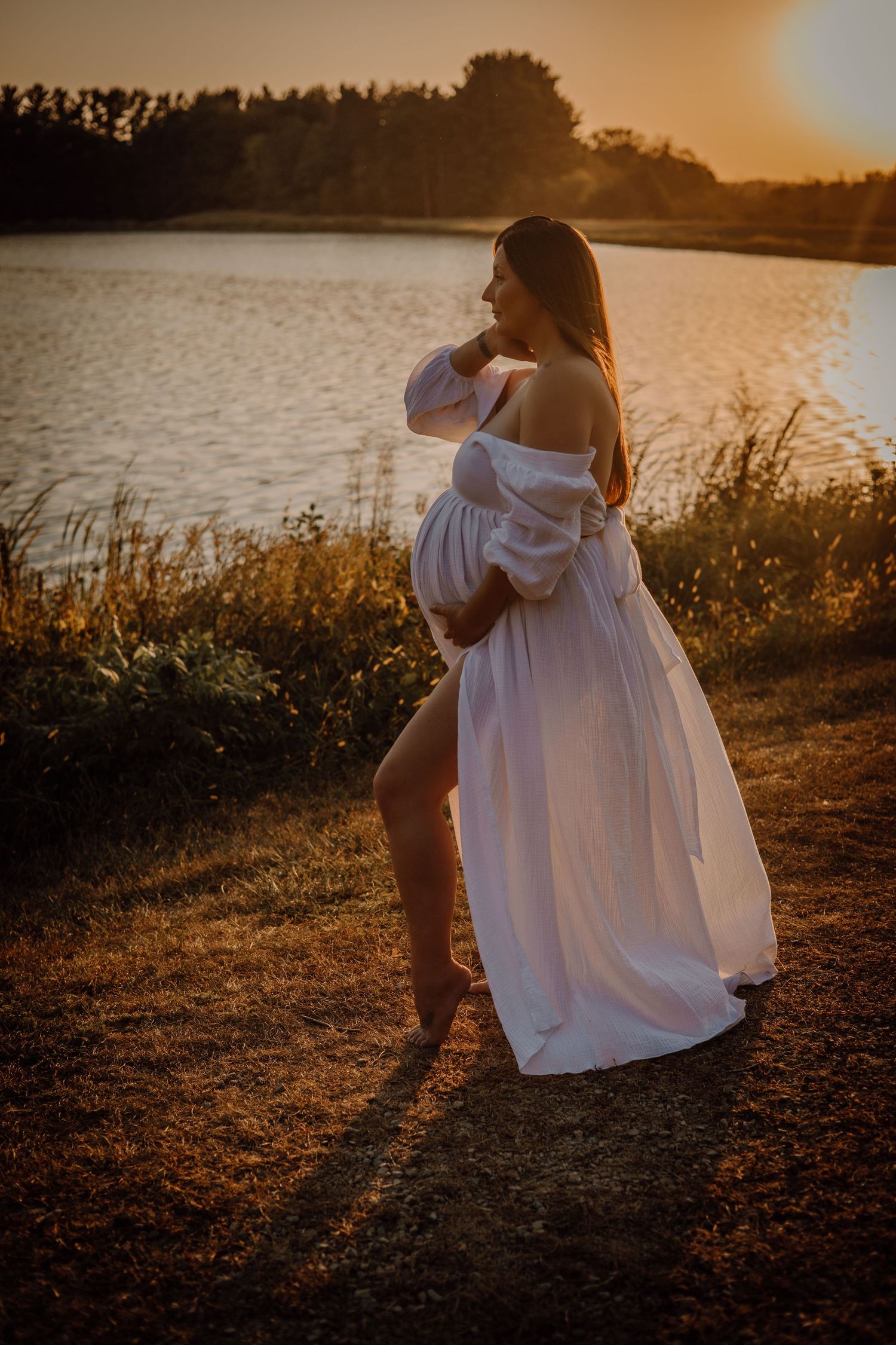 Person in flowing white dress sits by lakeside during golden sunset hour creating dreamy silhouette.