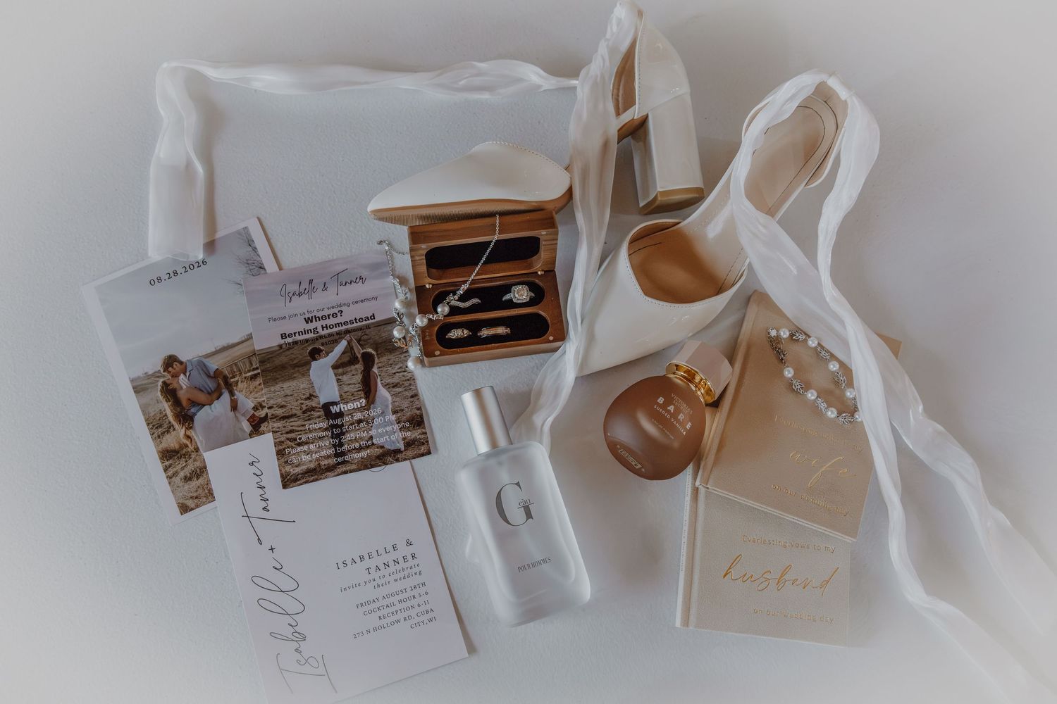 Elegant bridal flat lay with wedding rings, perfume, heels, pearls, and stationery on white fabric.