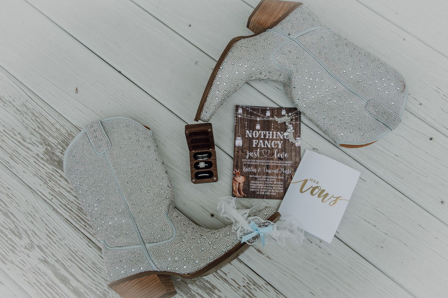 Glitter cowboy boots with wedding invitation, ring box, and vow booklet on white wood floor.