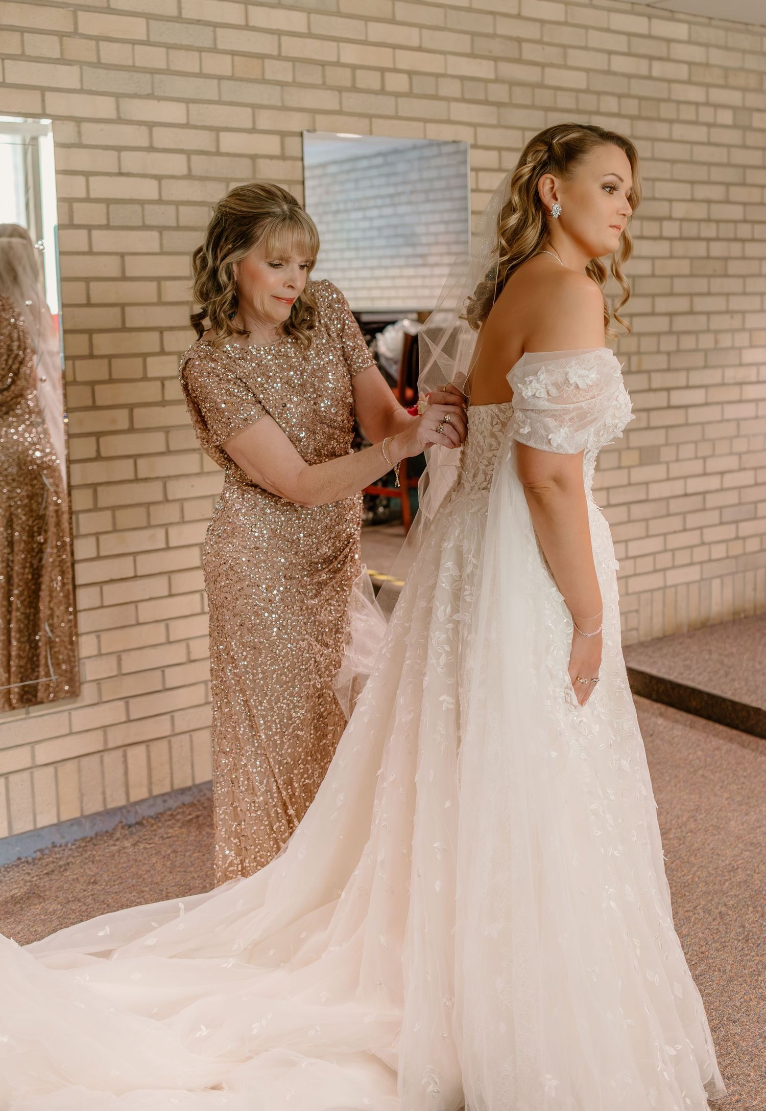 Helper adjusts the delicate lace train of an elegant off-shoulder wedding gown against a brick wall backdrop.