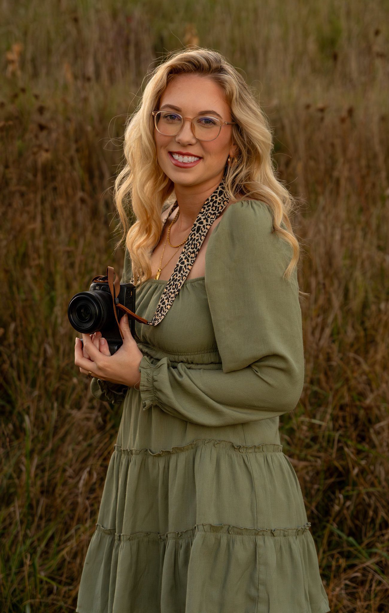 Professional photographer in olive green dress holds camera while posing in outdoor field setting.