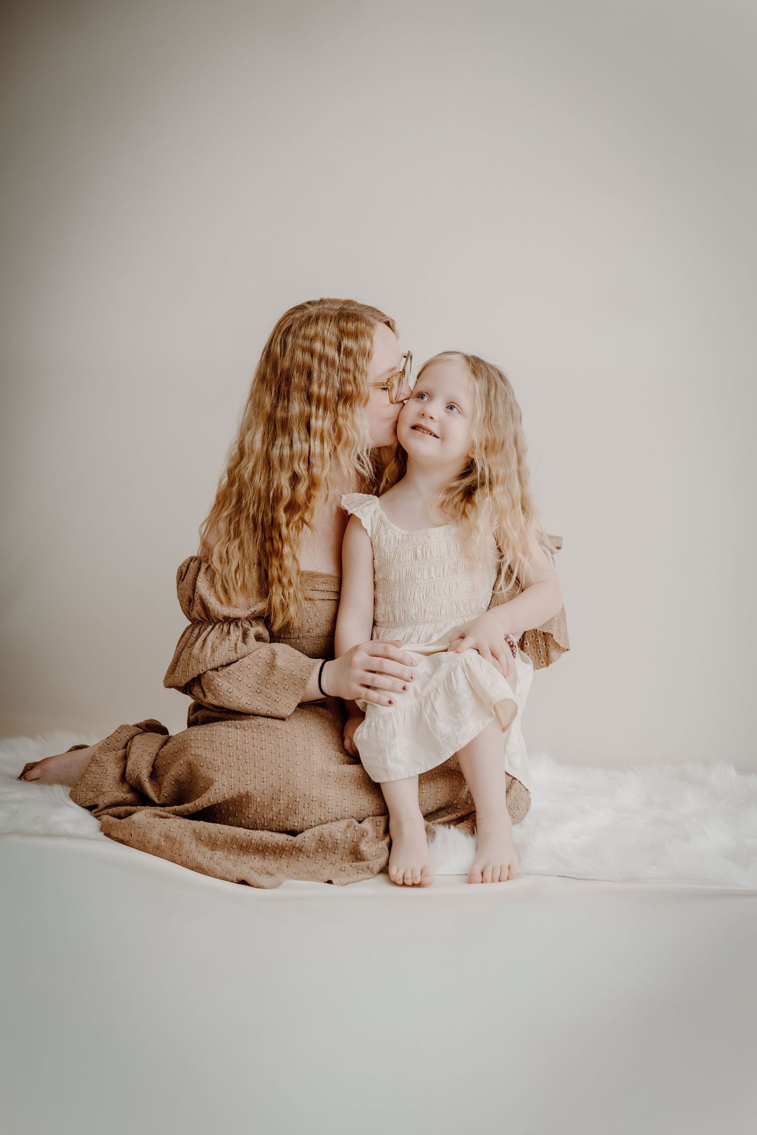 A mother with long hair kisses her young daughter's cheek as they sit together in matching neutral dresses.