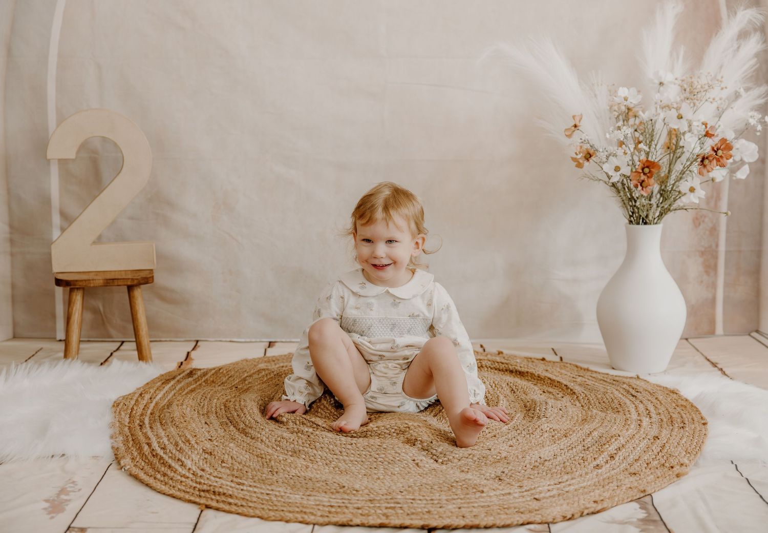 A baby in white clothing sits on a round jute rug next to a white vase with dried flowers in a neutral-toned setting.