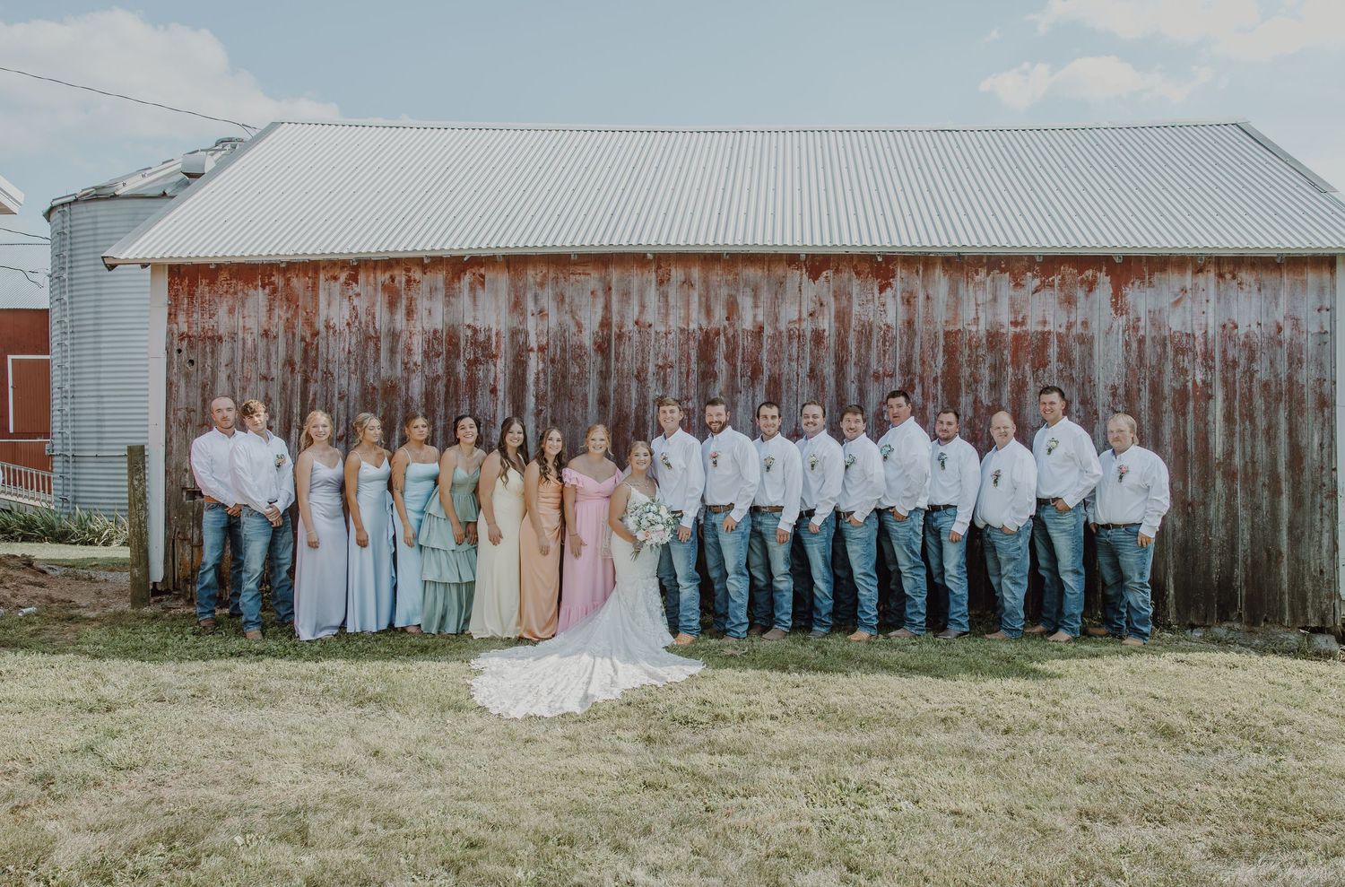 Wedding party poses in front of a rustic barn, bridesmaids in pastel gowns and groomsmen in jeans and white shirts.