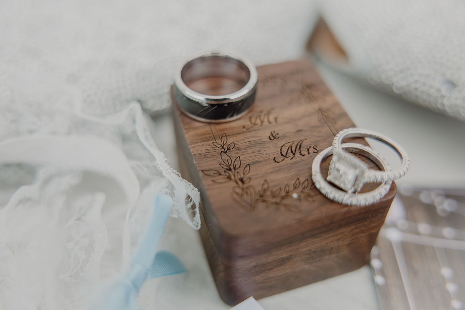 Wedding rings displayed on an engraved wooden Mr. & Mrs. ring box with lace and blue ribbon accents.