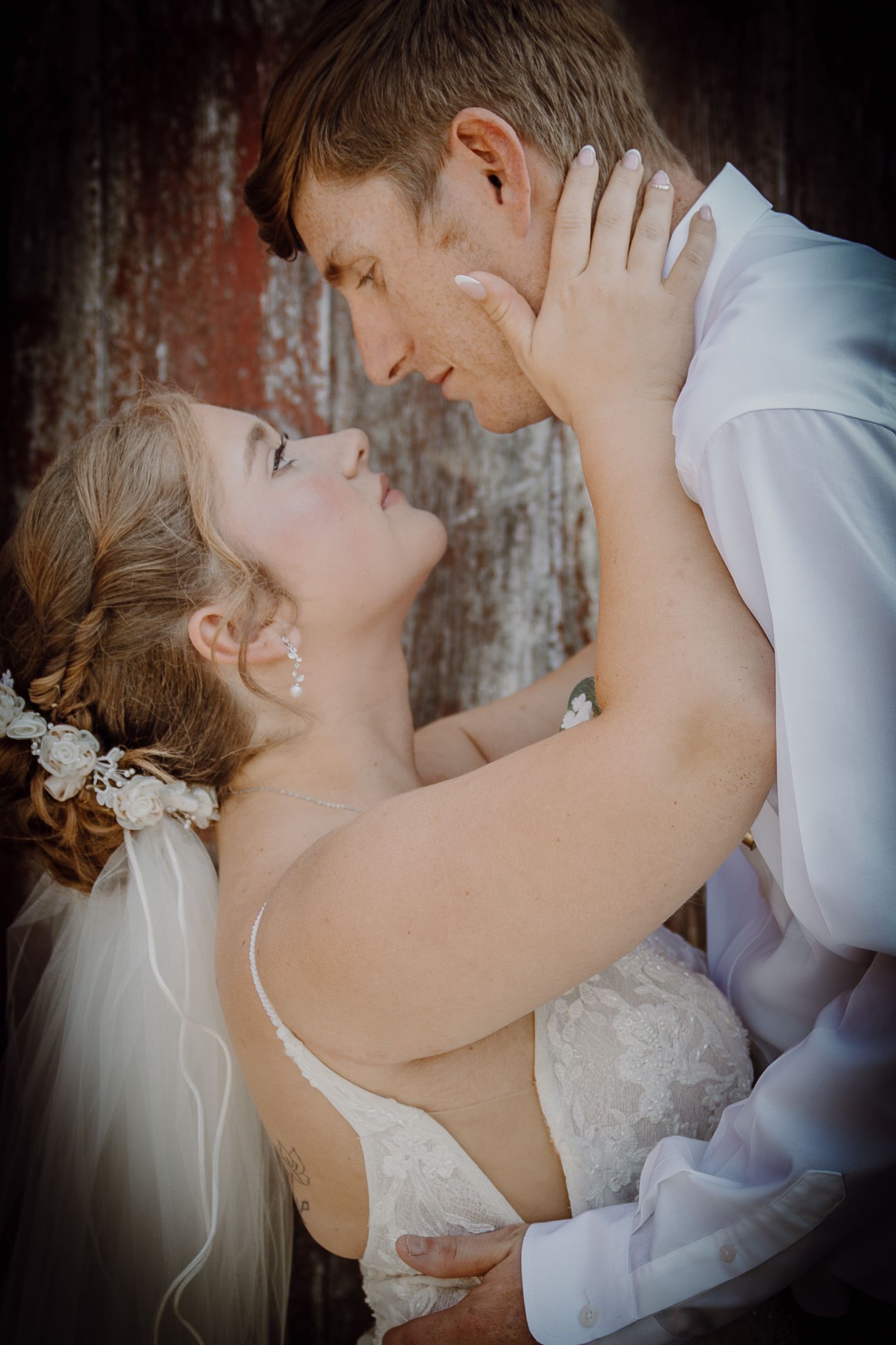 Bride and groom share a romantic moment, nearly kissing, with the bride in a lace gown and veil against a rustic backdrop.