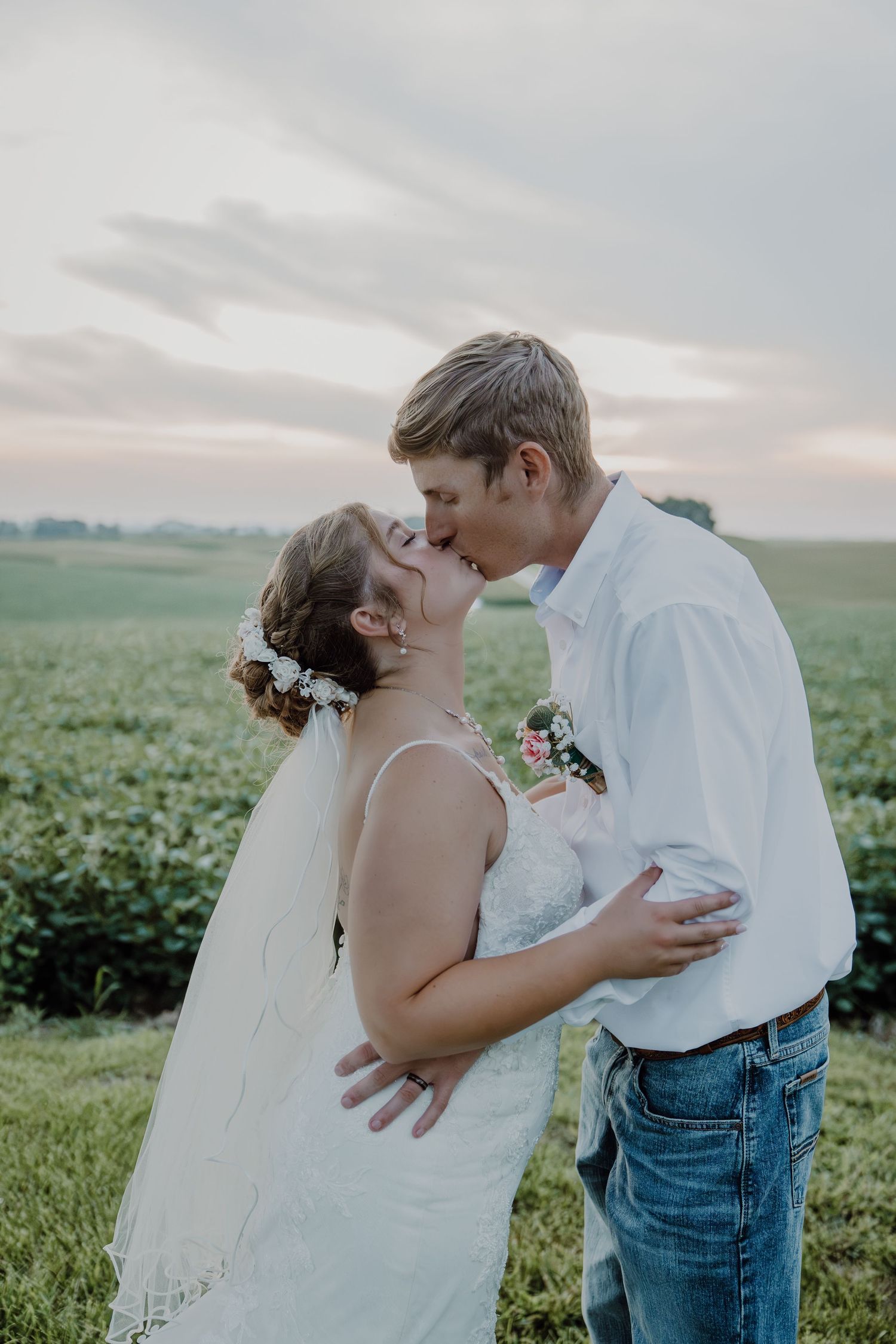 Bride and groom sharing a romantic kiss in a green field at sunset, bride wearing a white dress with floral hair piece.