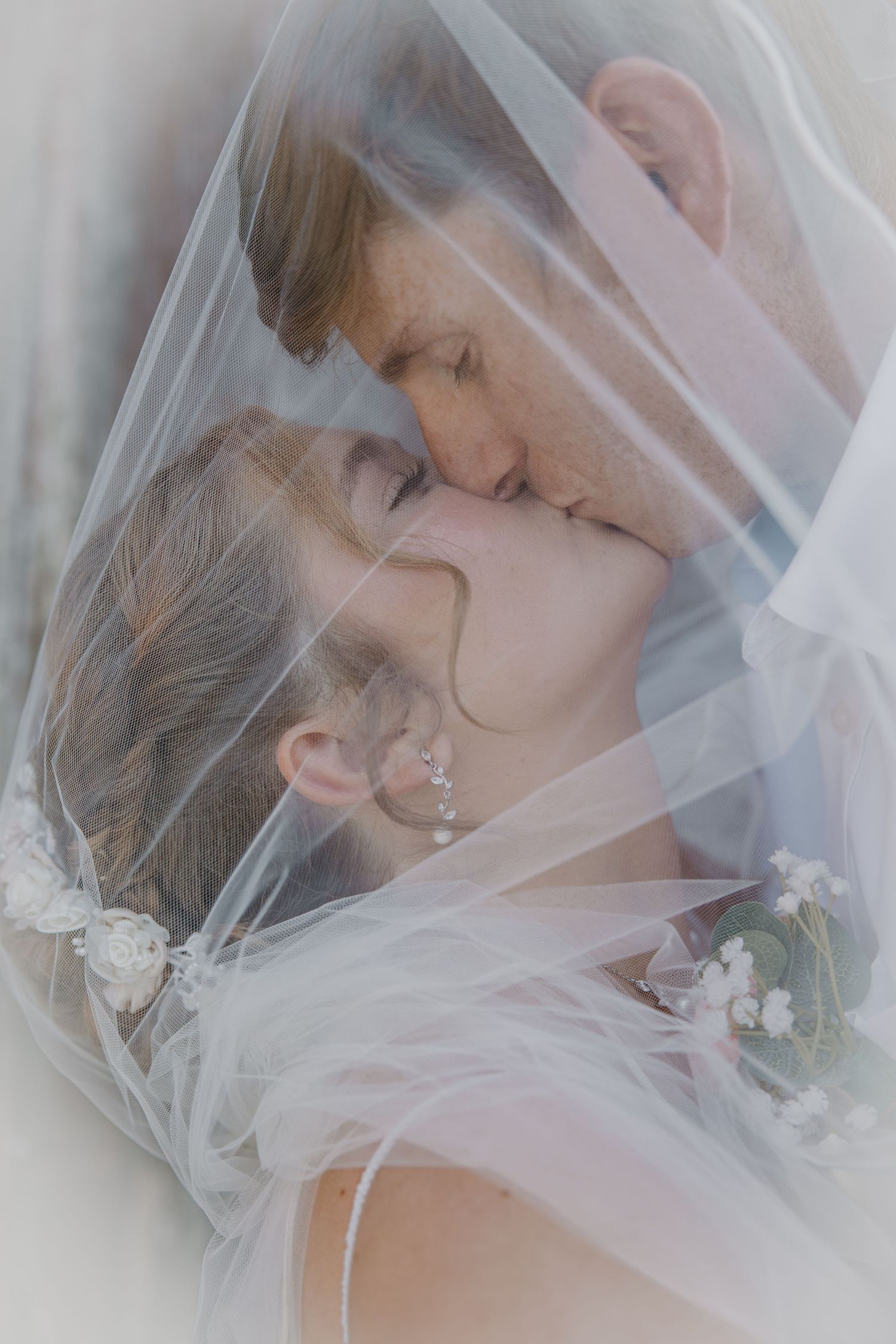 Bride and groom sharing a romantic kiss under a flowing white wedding veil, captured in a soft, dreamy close-up.