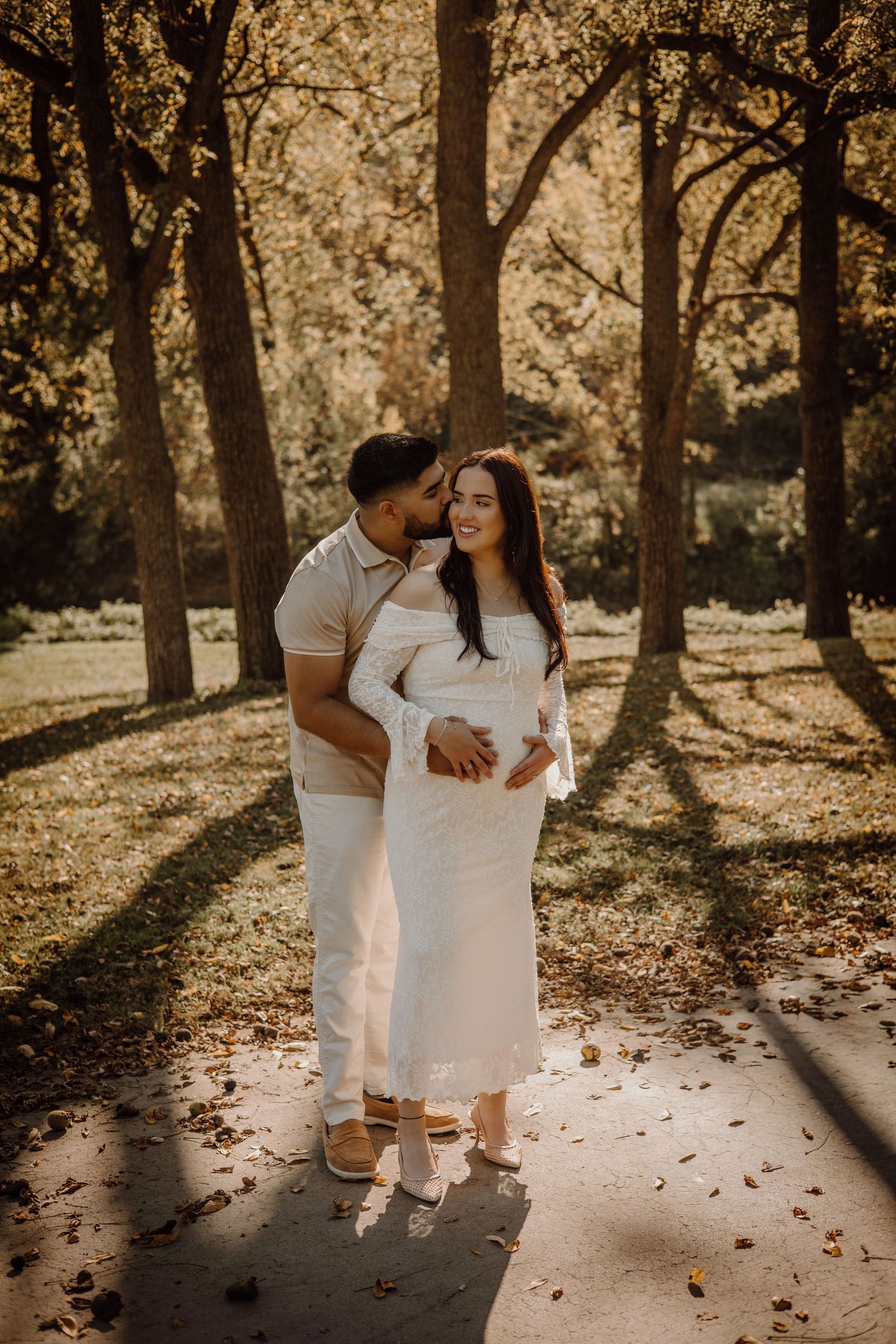 A couple in white outfits share an intimate moment in a sunlit forest with fallen leaves on the ground.