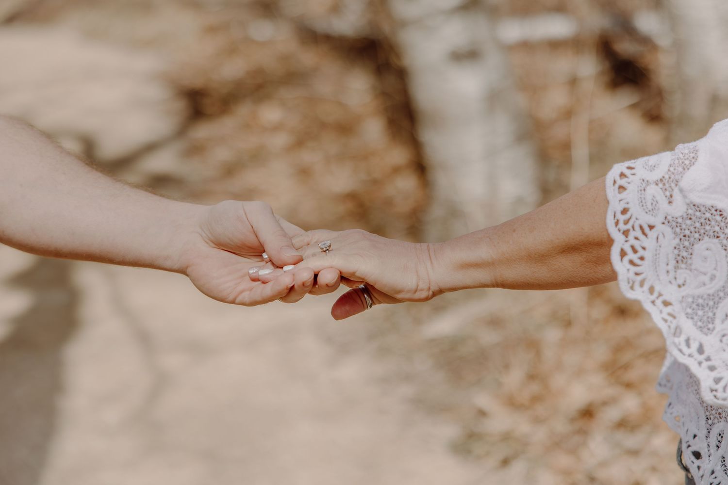 Close-up of two people holding hands outdoors, one wearing a wedding ring and lace dress sleeve.