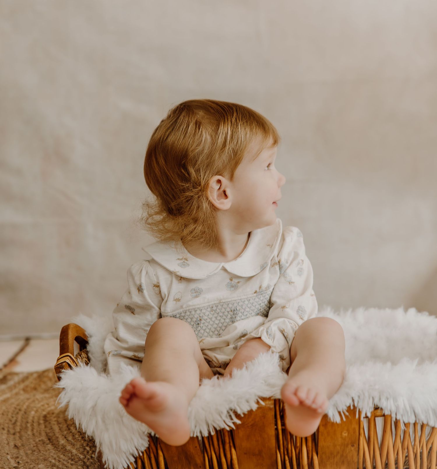 Child in white ruffled dress sits on woven basket against neutral background in artistic portrait series.