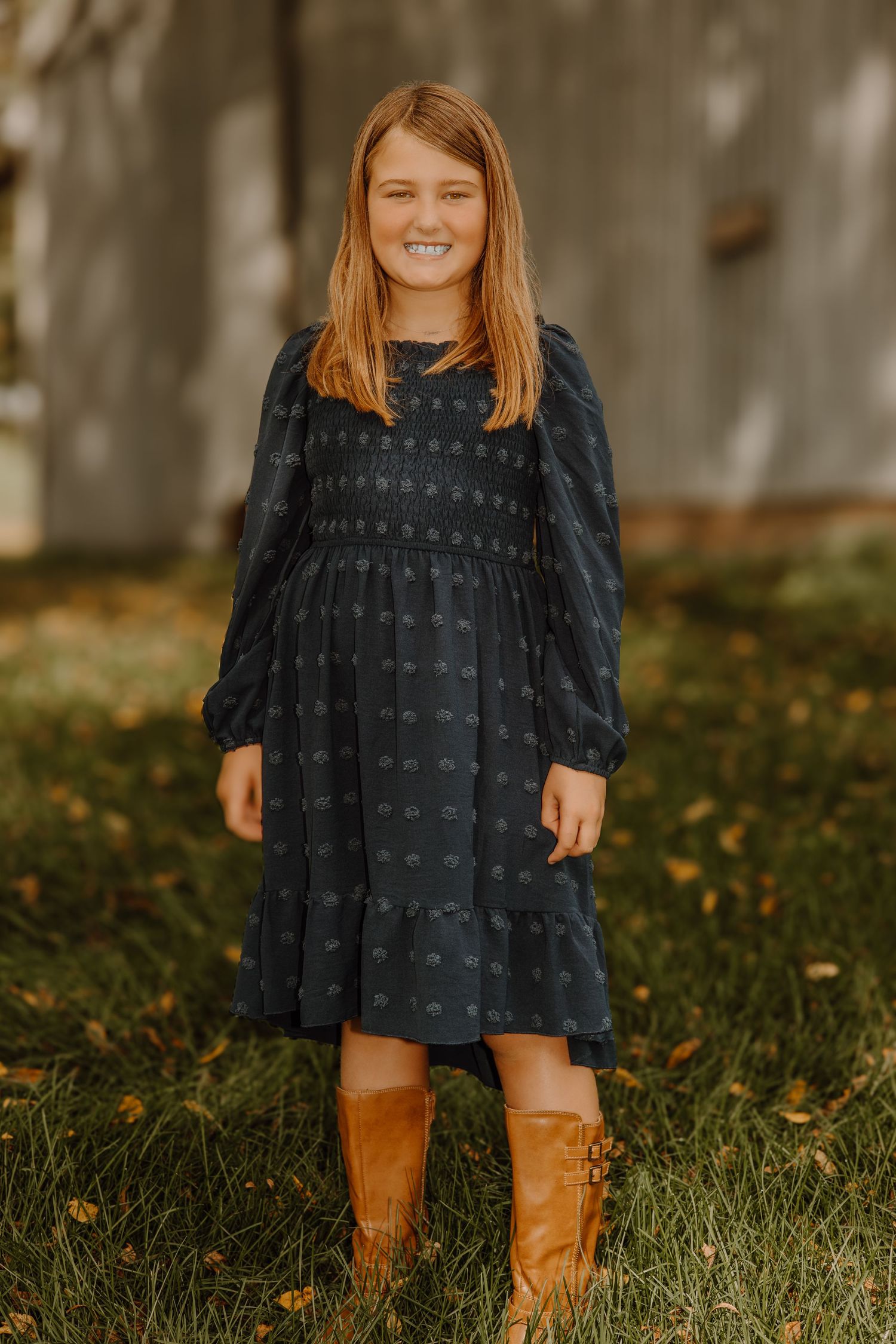 A child wearing a navy blue dress and brown boots stands in grass with fallen autumn leaves.