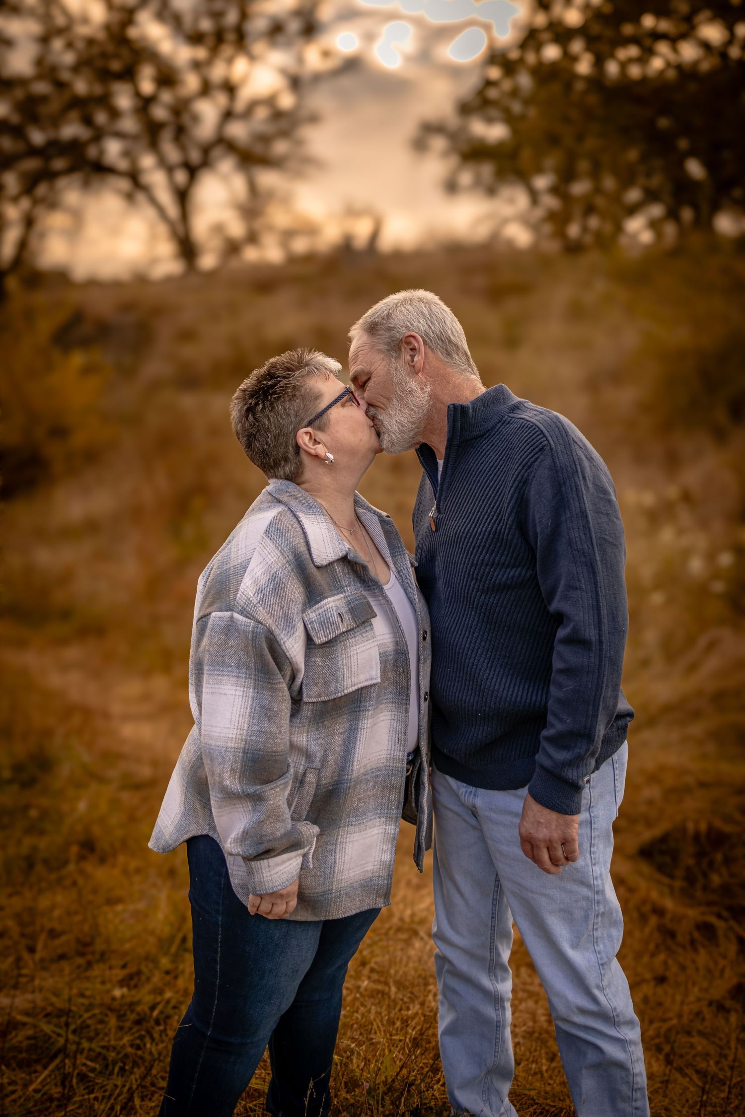 A couple shares a tender kiss in a field at sunset while wearing casual attire in a series of romantic photos.