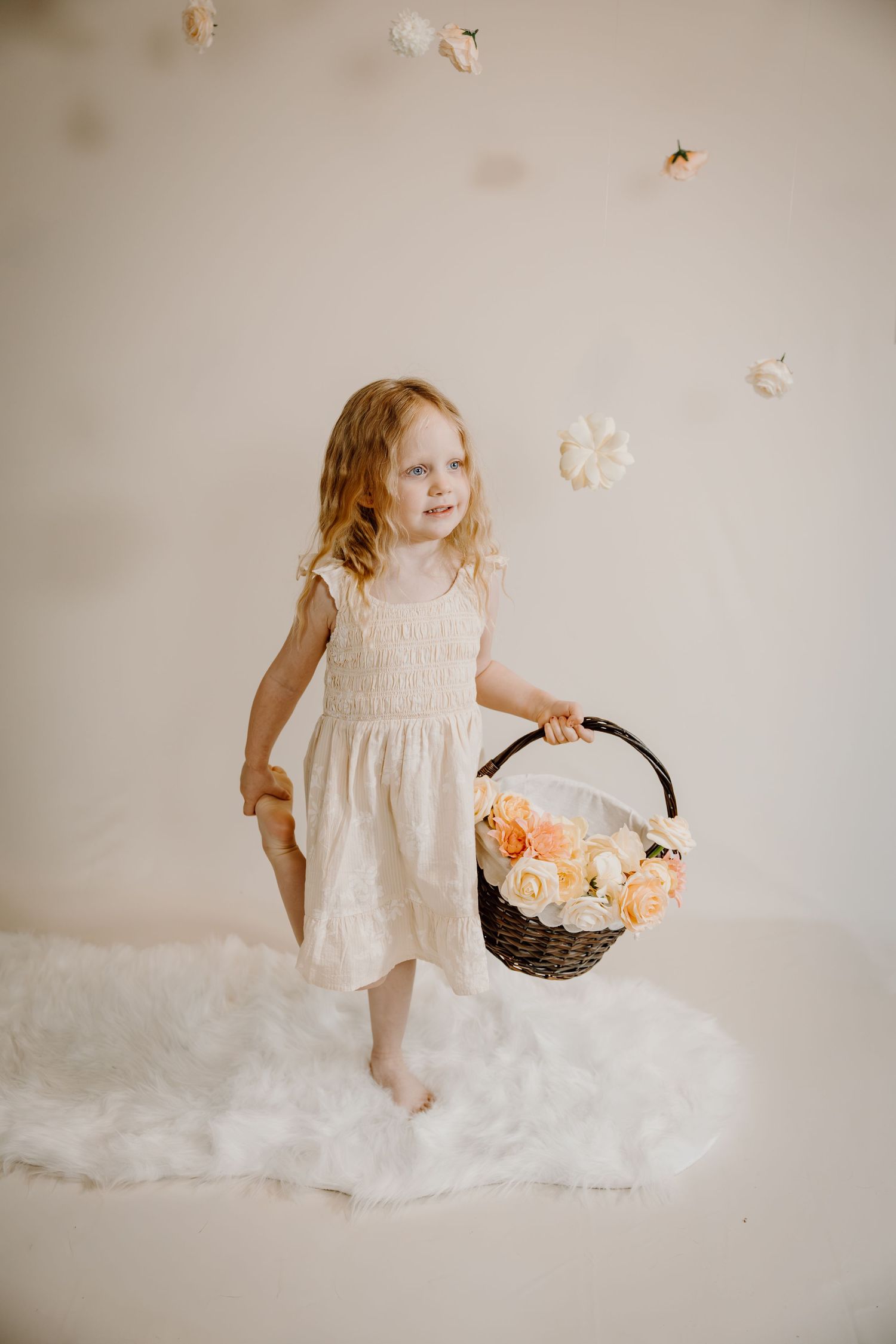Young girl in white dress holding flower basket with roses, standing on fluffy rug against neutral backdrop.