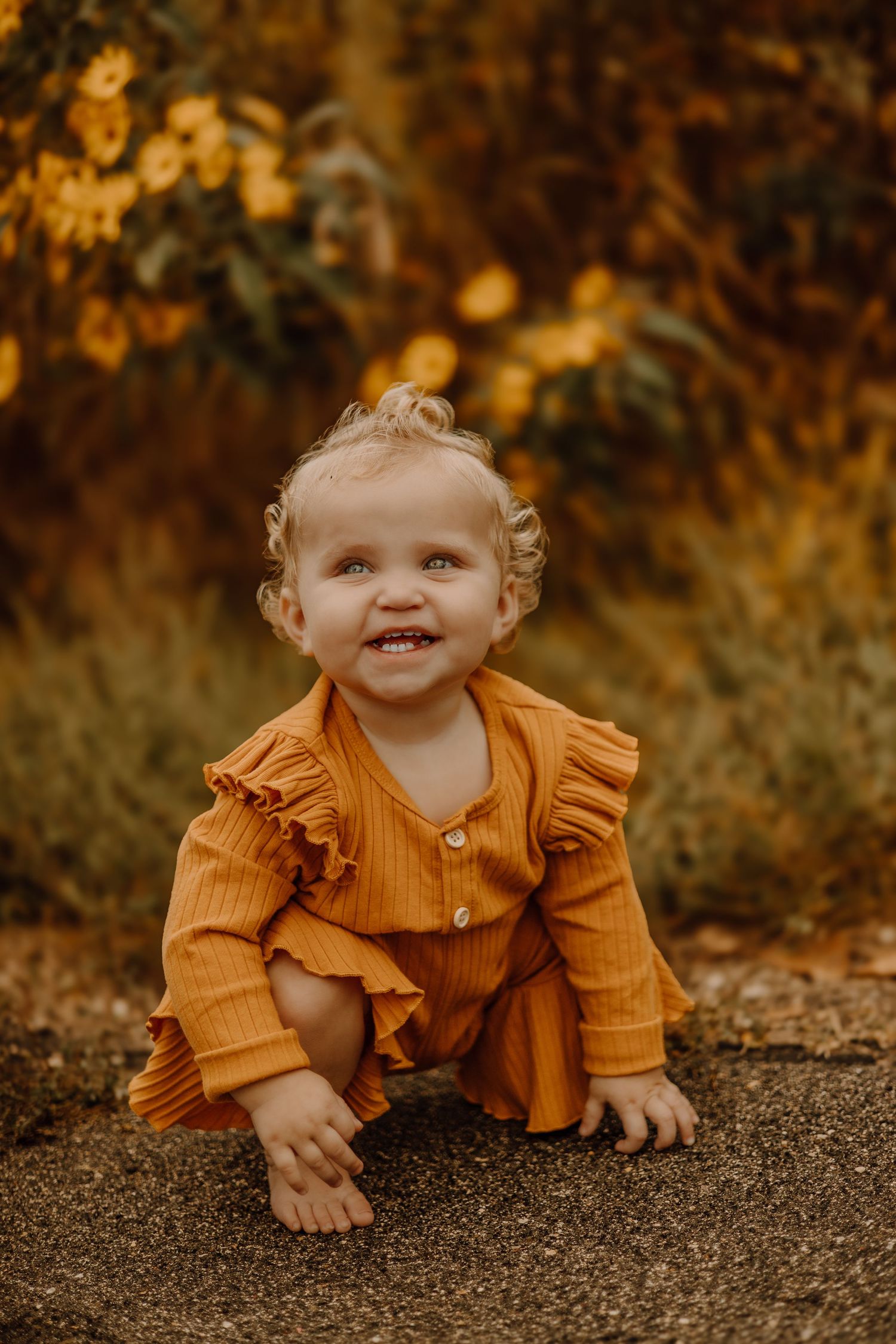 Small child in mustard yellow ruffled outfit crawls and smiles on ground during autumn photo session.