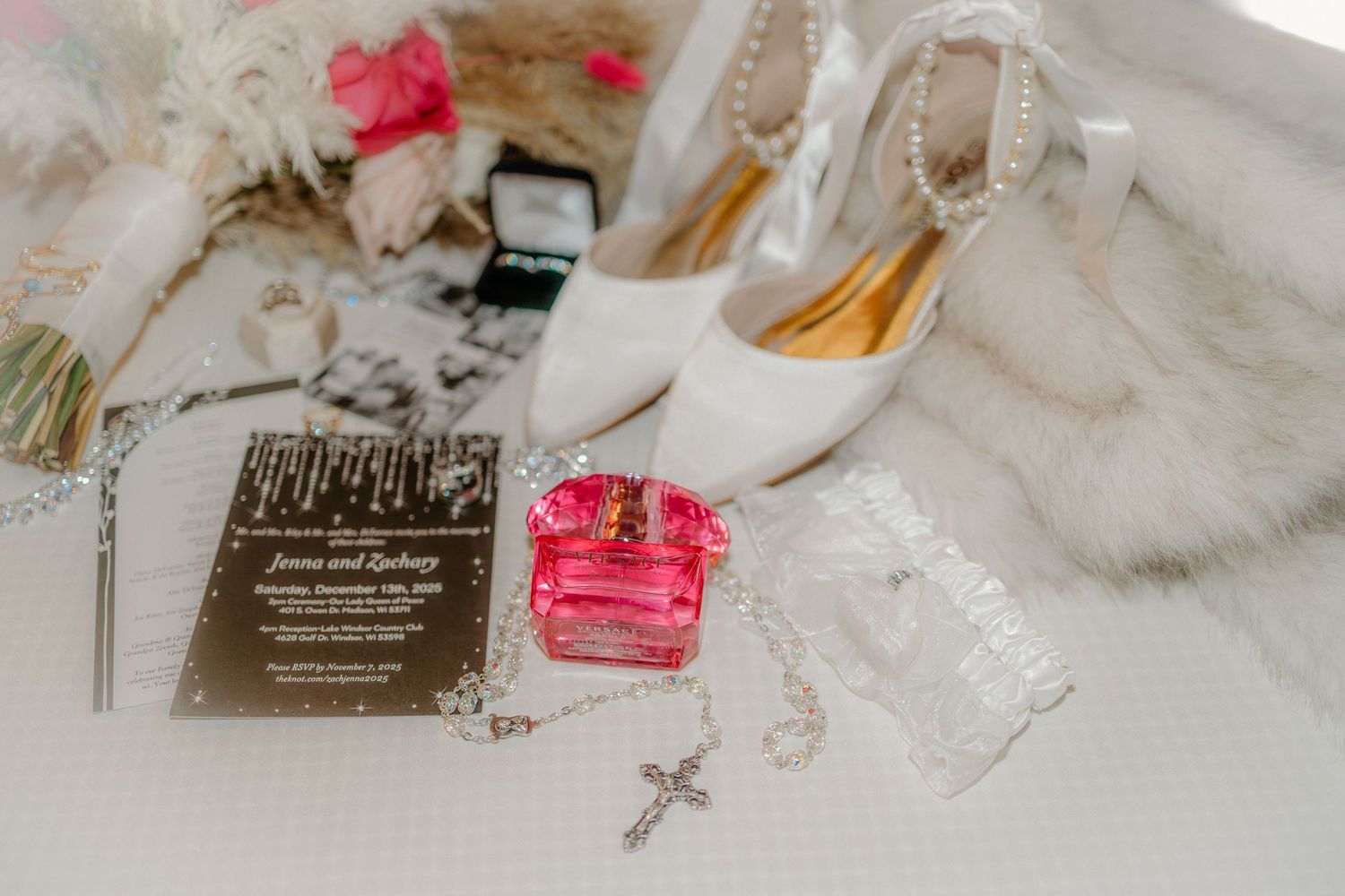 Wedding details including pink floral elements, invitations, rings, and religious cross laid out on white fabric.
