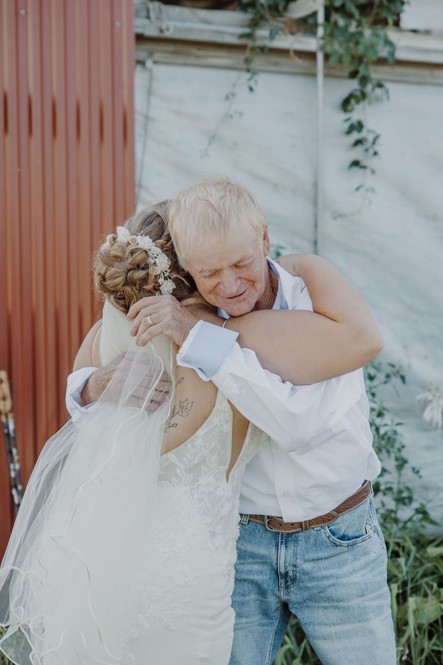 Emotional father-daughter wedding hug, bride in white gown and veil, older man in white shirt and jeans, outdoor ceremony.