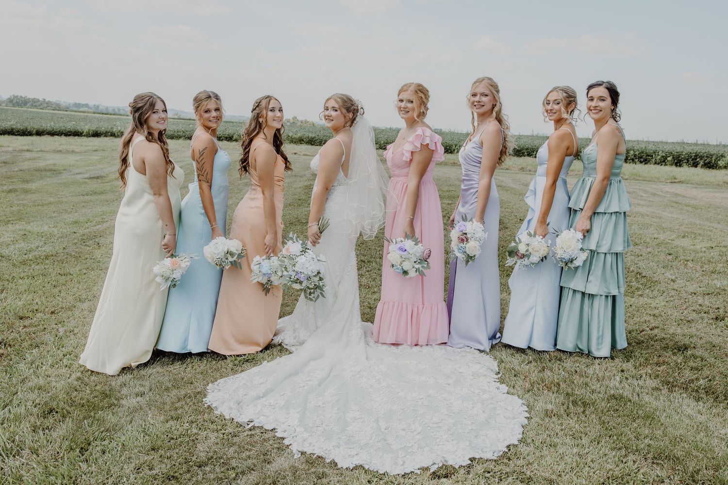 Bride with bridesmaids in pastel mismatched gowns holding white bouquets on a green field at an outdoor wedding.
