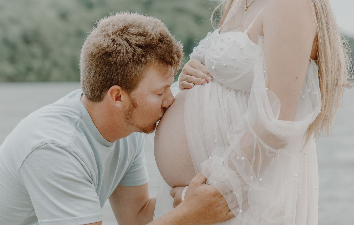 A man kneels kissing a pregnant woman's bare belly during an outdoor maternity photo session.
