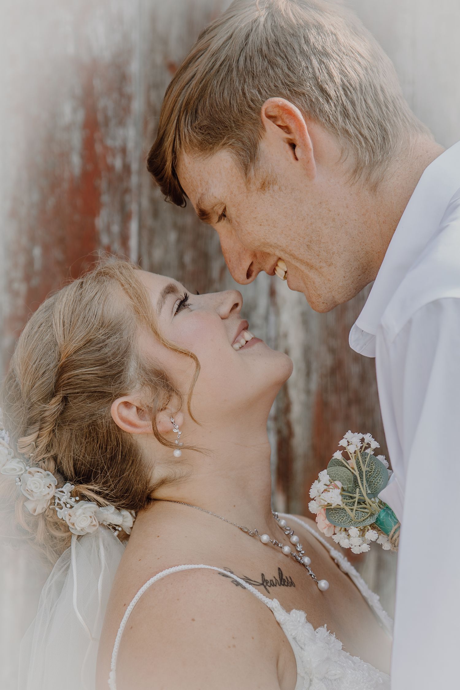 A smiling bride and groom share a romantic moment, nearly kissing, with wedding florals and rustic background.