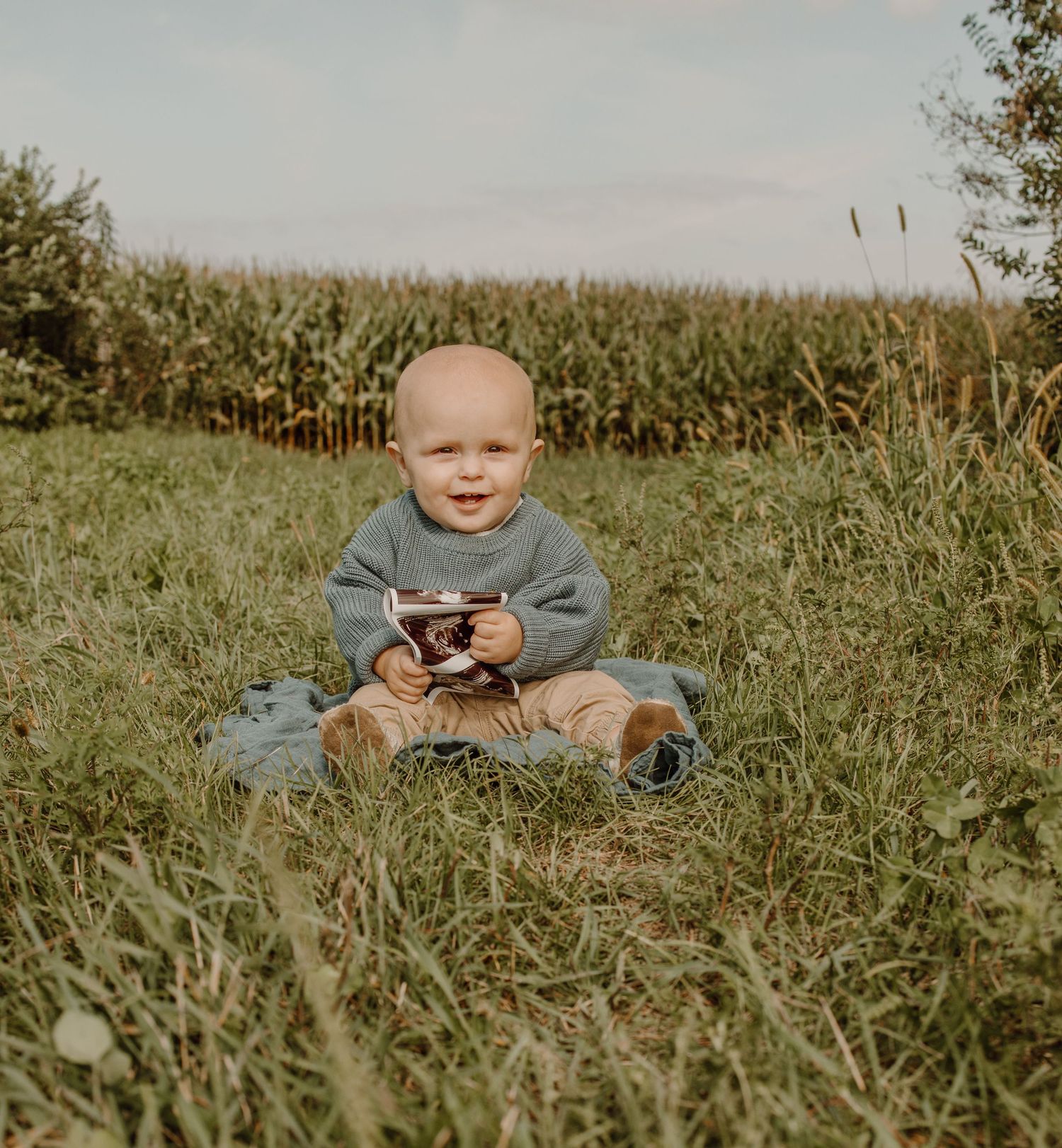 Small child sits in green grass holding a vintage camera with cornfield in background.