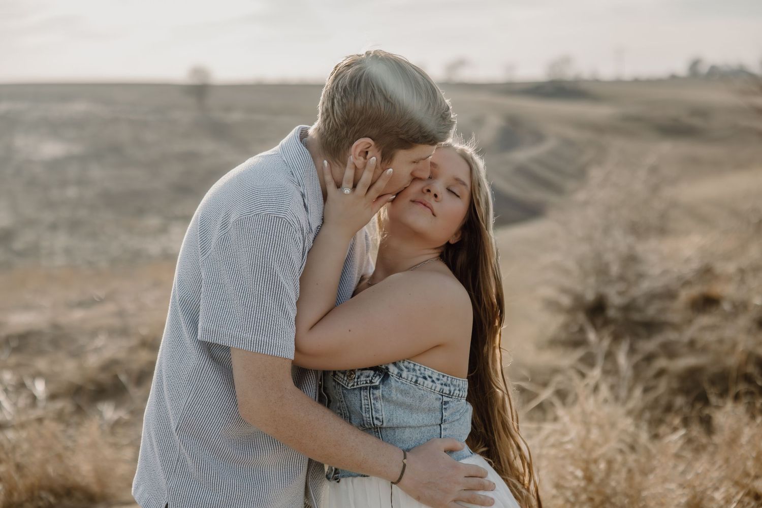 Couple embraces tenderly in a romantic outdoor photoshoot against a scenic mountainous backdrop at sunset.