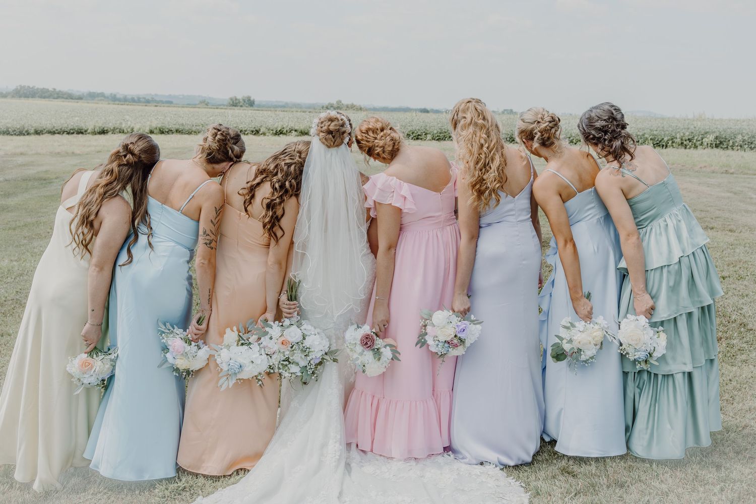 Bride and bridesmaids in pastel mismatched gowns holding bouquets, posing with backs to camera in a field.