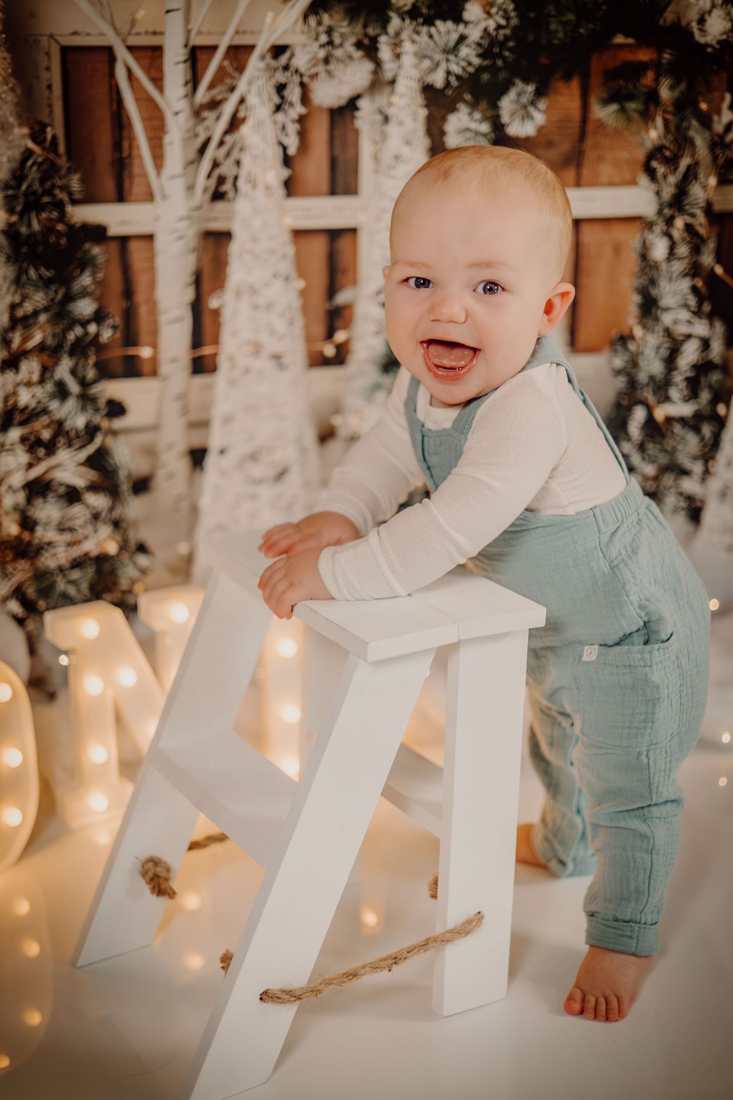 Baby in white sweater and blue pants stands by illuminated letter decor with twinkling lights in cozy winter scene.