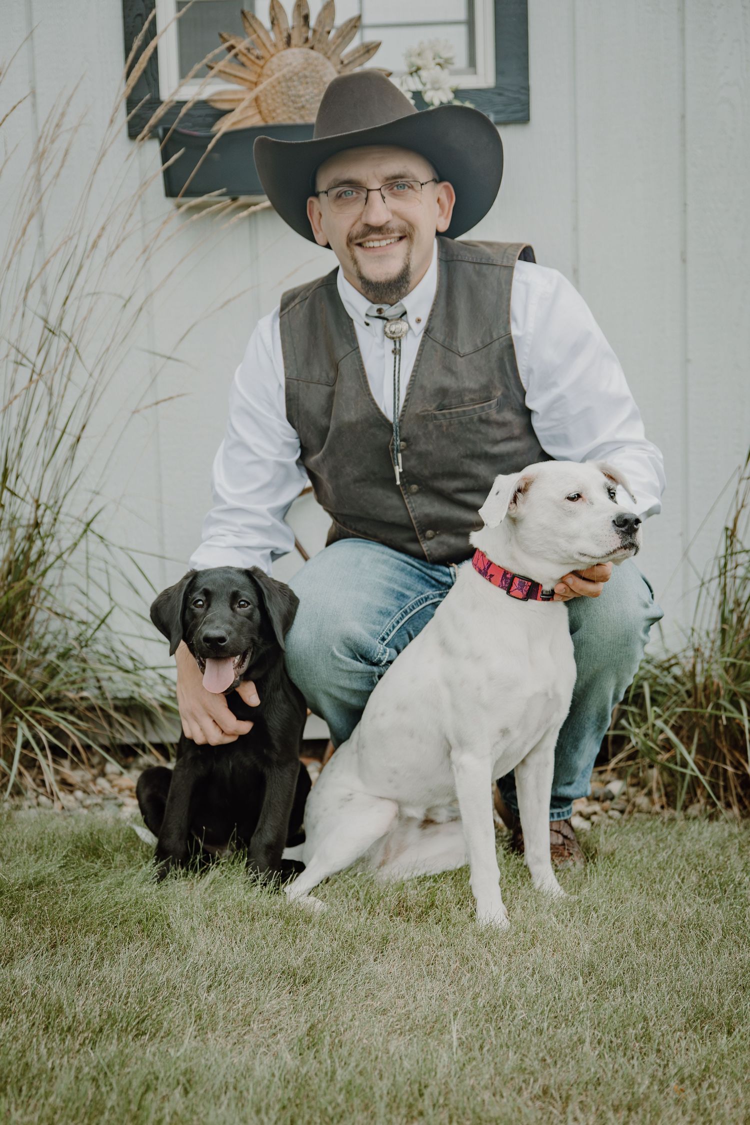 Man in cowboy hat and vest kneeling with black Labrador puppy and white dog with red collar outdoors.