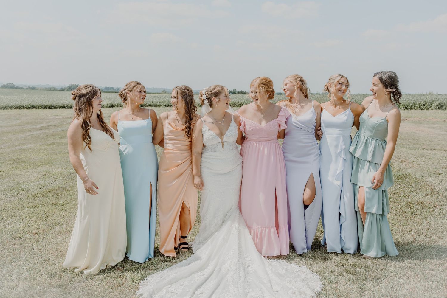Bride in white gown poses with seven bridesmaids in pastel dresses outdoors on a sunny wedding day.
