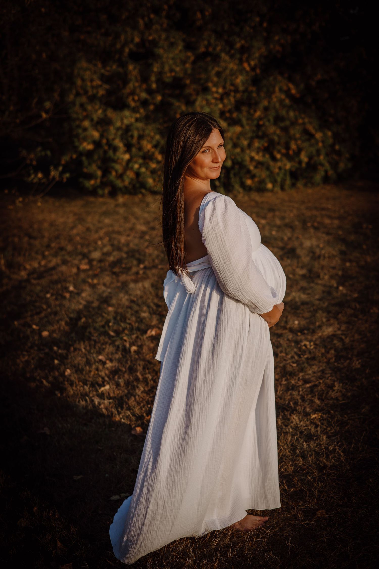 A woman in a flowing white dress poses in a natural outdoor setting during golden hour.
