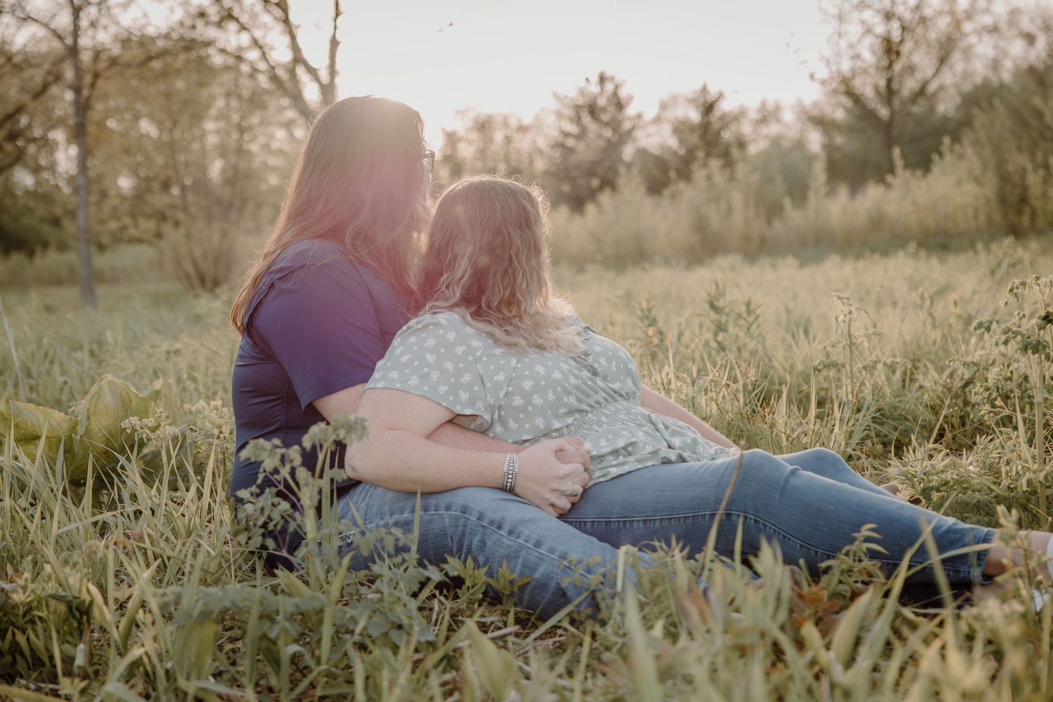 Two women sitting together in a sunlit meadow, sharing a tender moment surrounded by tall grass and trees.