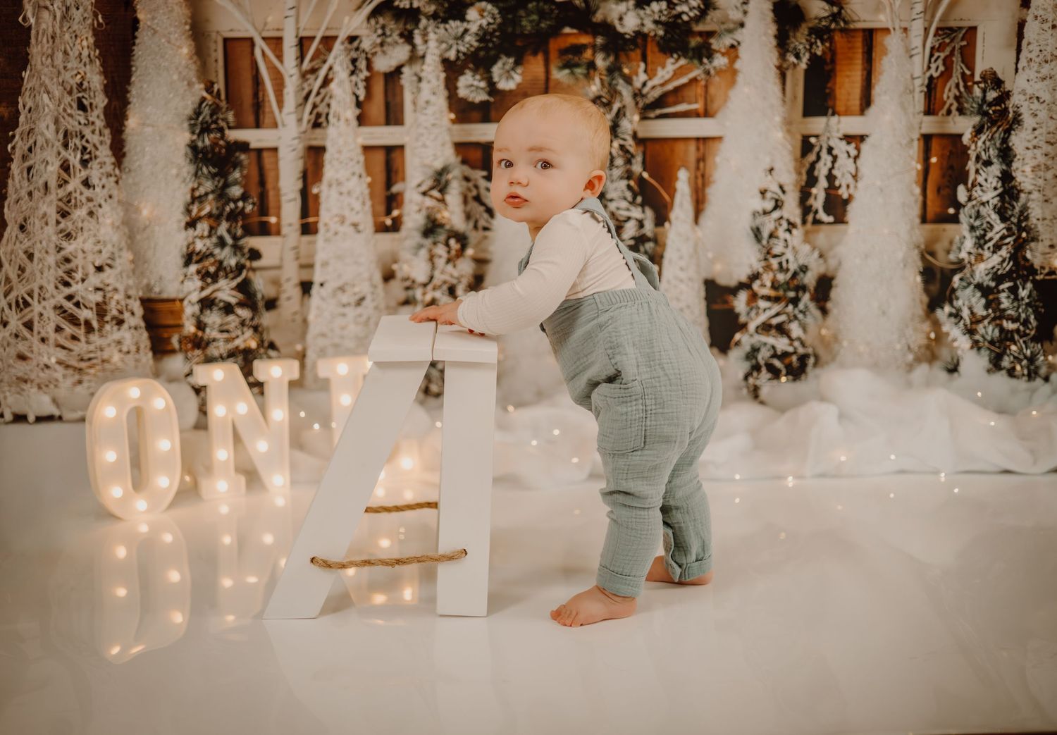 A small child in overalls stands near a decorative winter wonderland setting with twinkling lights and snow.