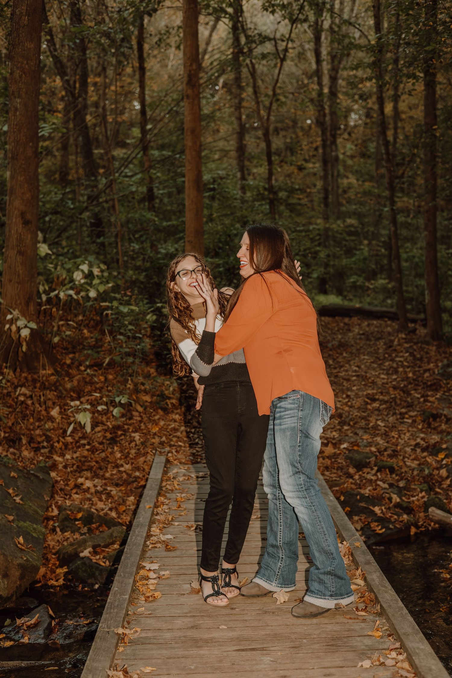 A couple shares a romantic moment on a wooden boardwalk trail surrounded by autumn trees and fallen leaves.