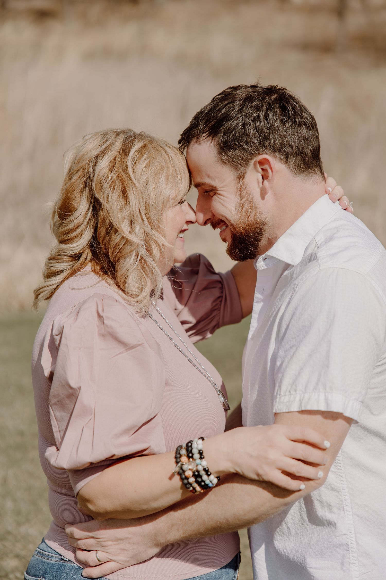 A smiling couple in casual attire share a romantic forehead touch outdoors, the woman wearing bracelets and a pink top.