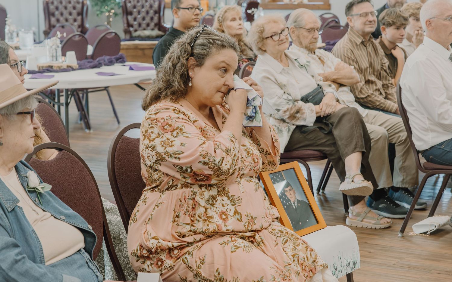 Emotional woman in floral dress holds framed photo at a memorial gathering with seated attendees in background.