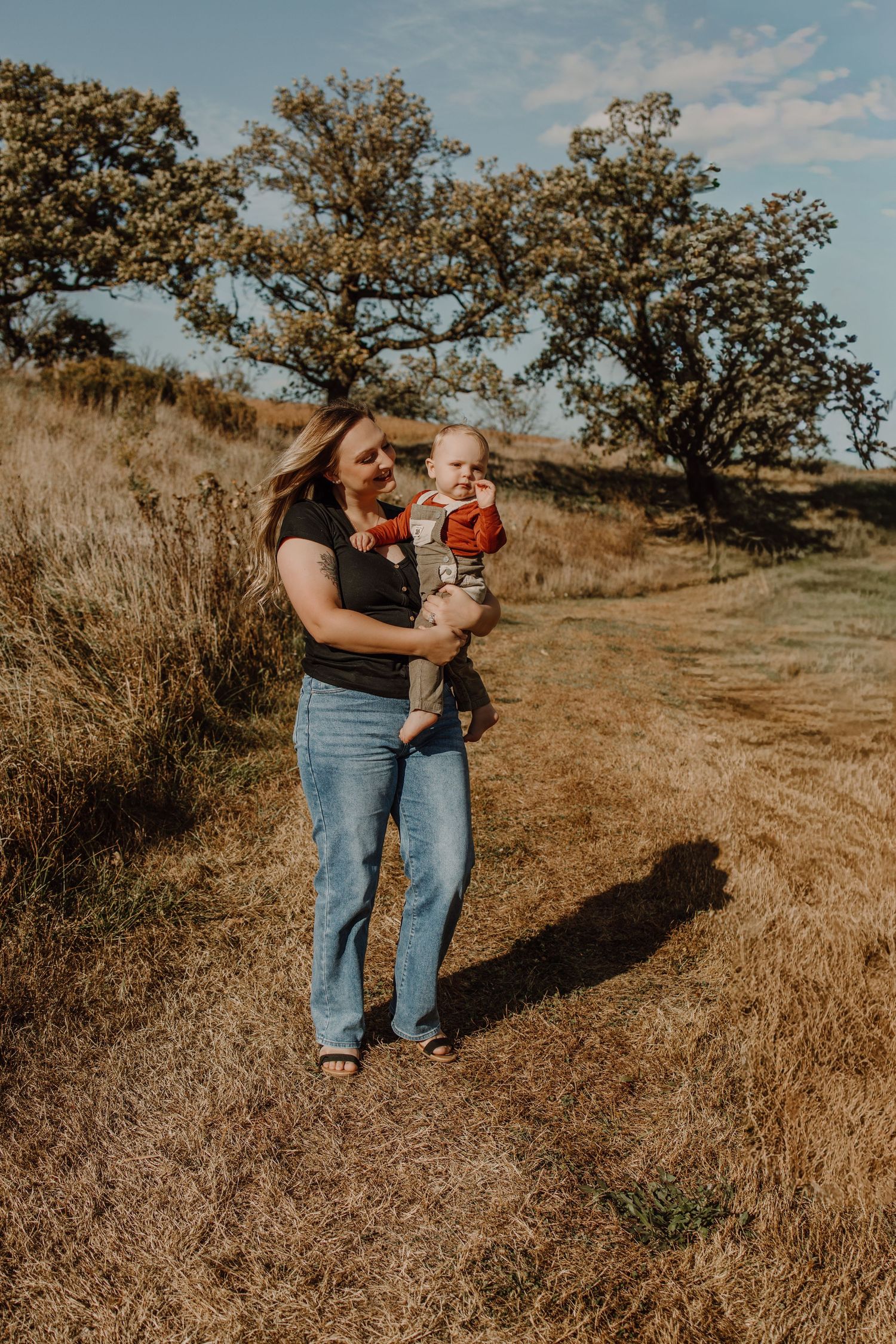 A parent and child walk along a grassy hillside trail on a sunny autumn day with trees in the background.